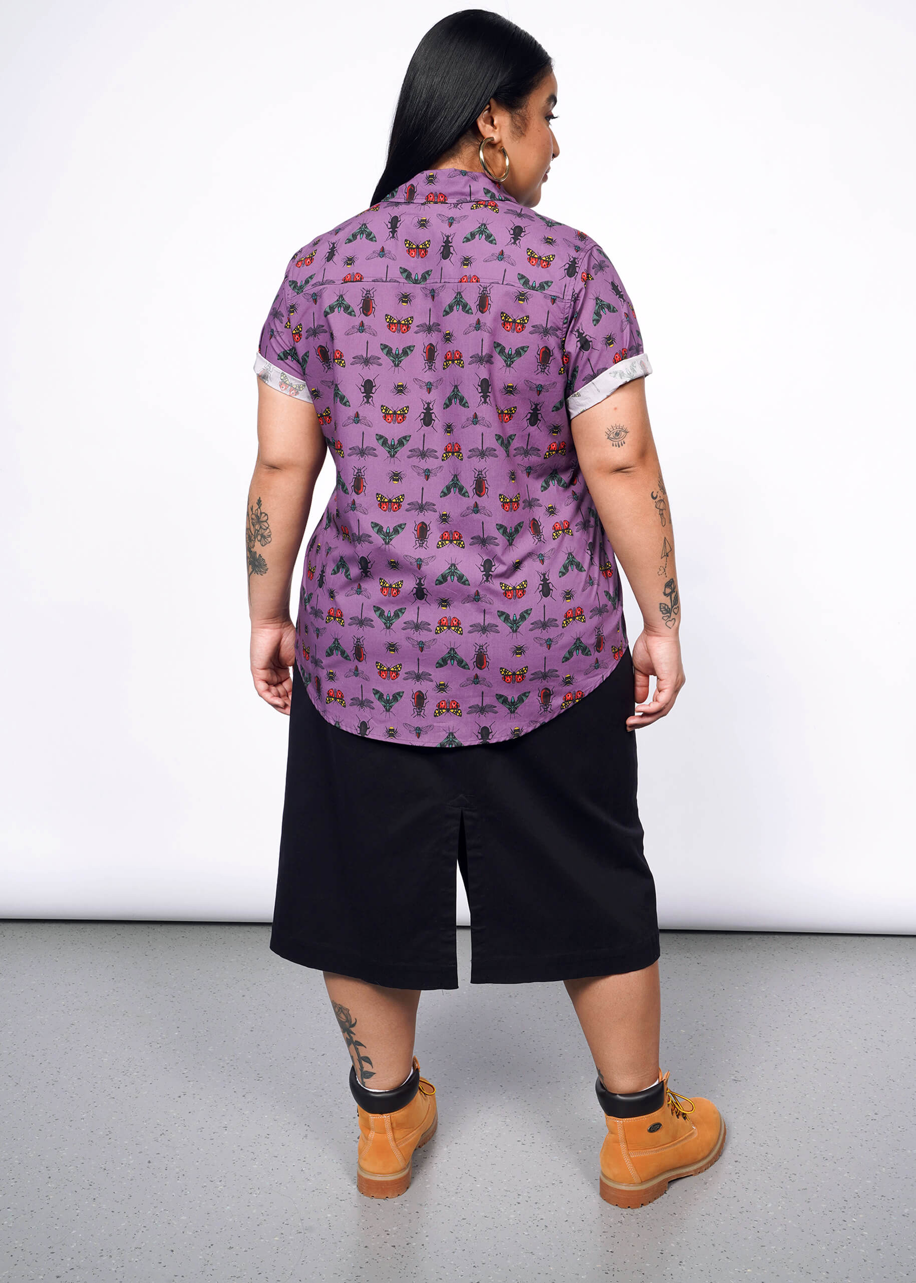 Back view of person with long black hair wearing hoop earrings, a purple bug printed button up, black wide leg shorts, and timberland boots.