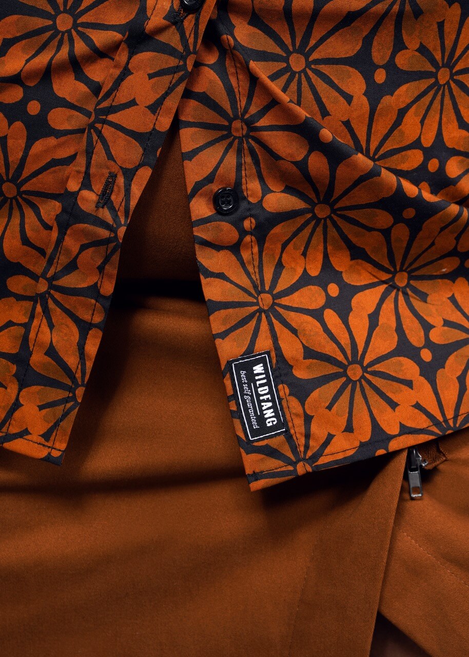 A close-up shows someone in The Essential Button Up by Wildfang, featuring a tailored fit with a bold orange floral pattern on black. Made from breathable cotton, its stylishly paired with matching dark orange attire.