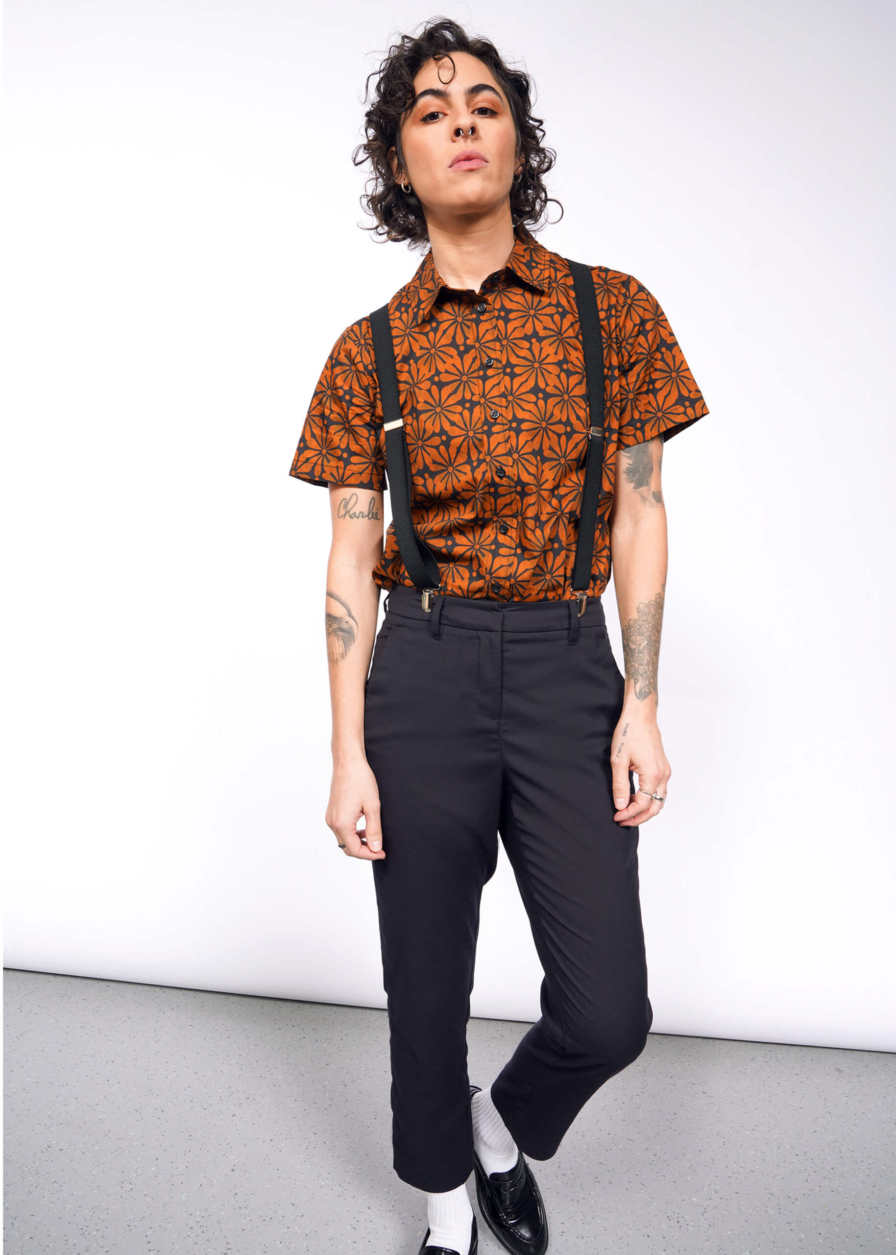 A curly-haired person in The Essential Button Up, an orange patterned, tailored-fit shirt, styled with black suspenders and pants, stands with one foot forward. Tattoos adorn their arms as they pose in a minimalistic indoor space.