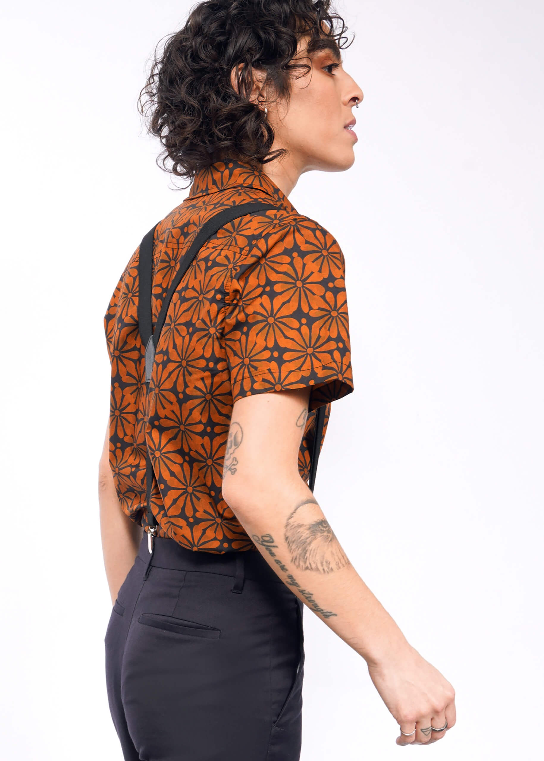 A person with curly hair displays a tailored fit, wearing The Essential Button Up in an orange and black pattern, paired with suspenders and black pants. Tattoos grace their left arm as they pose sideways against a plain background.