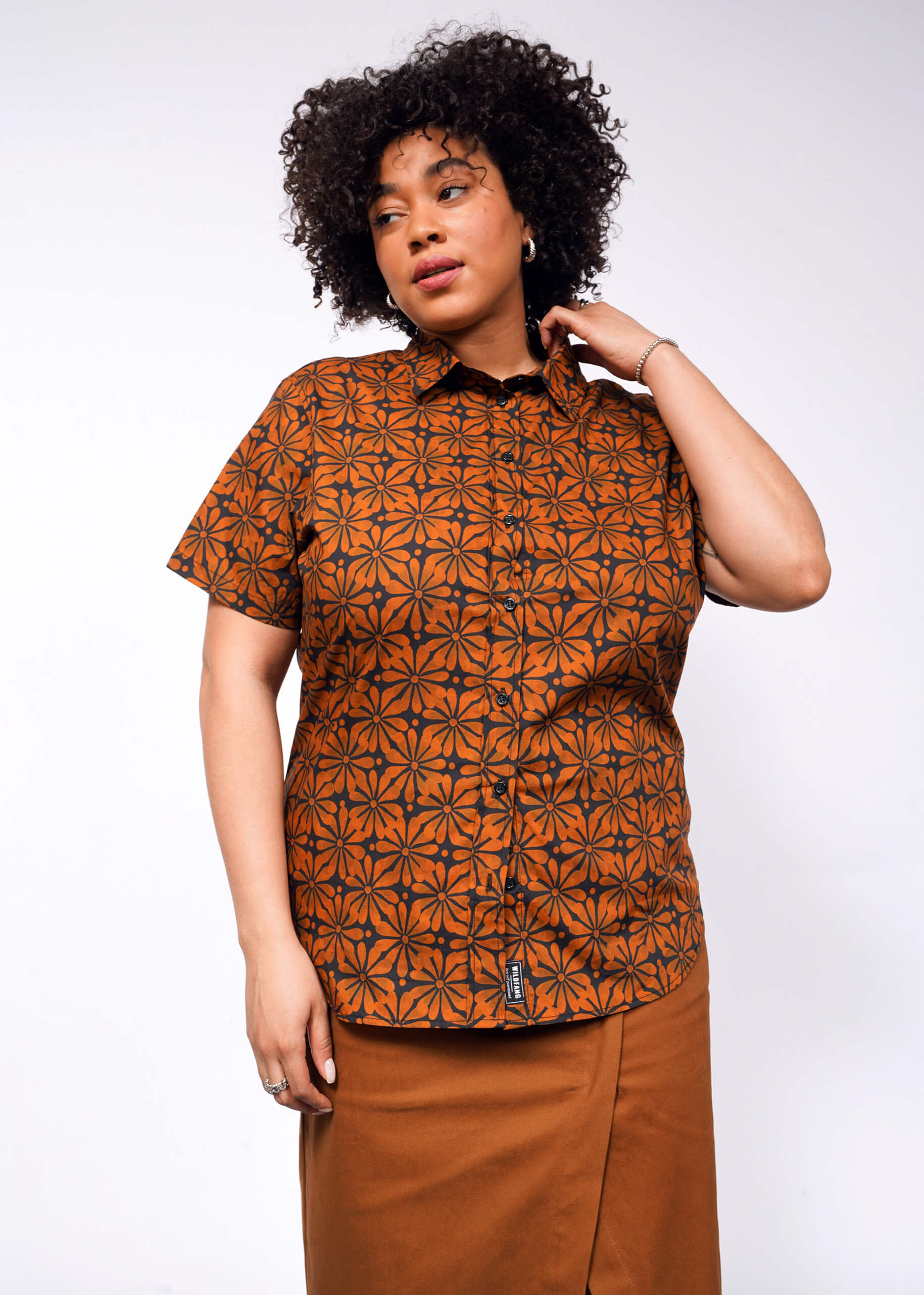 A woman with curly hair wears The Essential Button Up, a tailored-fit, patterned orange shirt, paired with a brown skirt. She stands against a white background, gazing to the side with one hand near her neck.