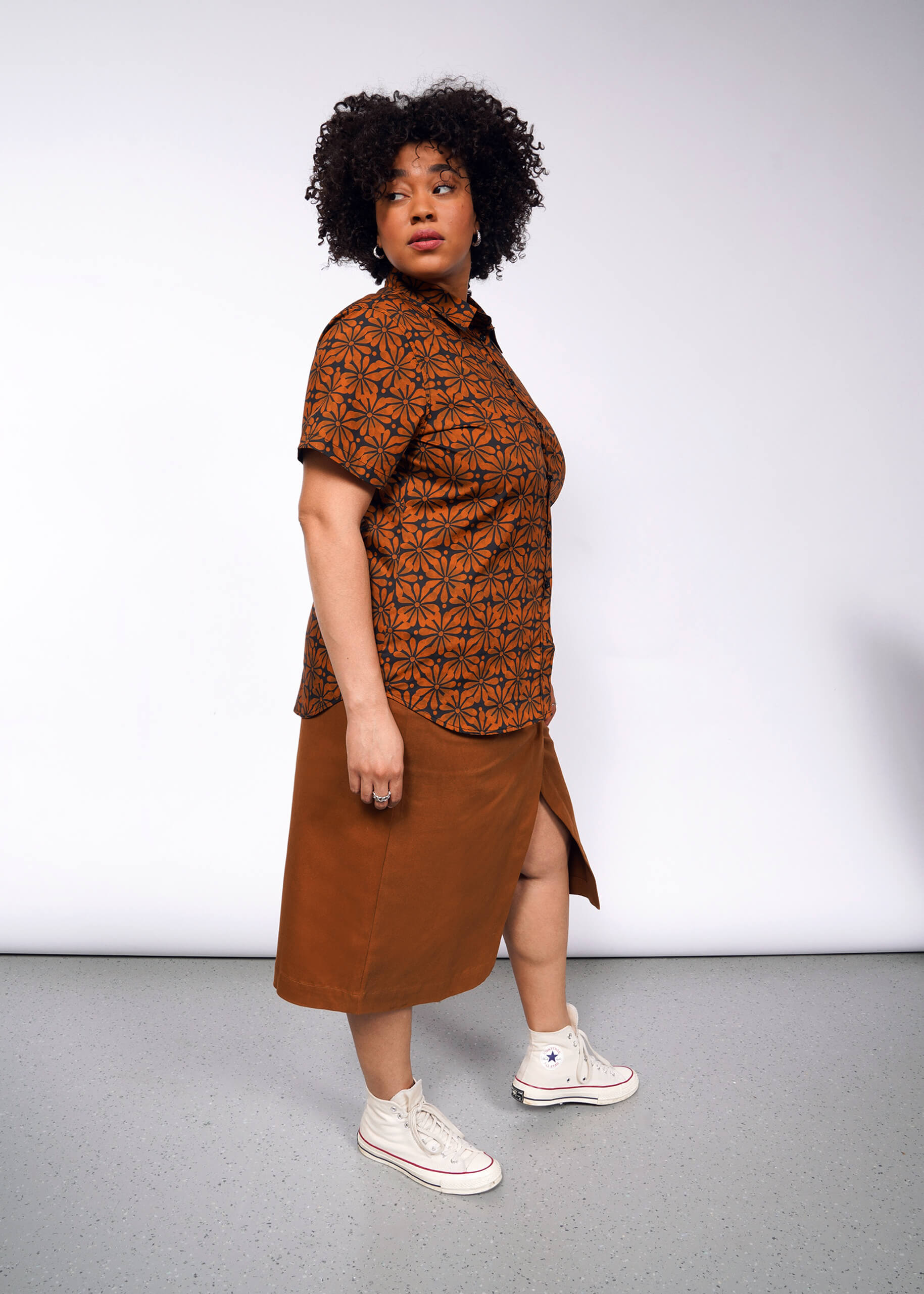 Wearing The Essential Button Up, a person with curly hair confidently poses in a brown patterned short-sleeve button-up shirt and brown skirt with a side slit, both made from breathable cotton, against a plain gray background. They complete their look with white high-top sneakers.