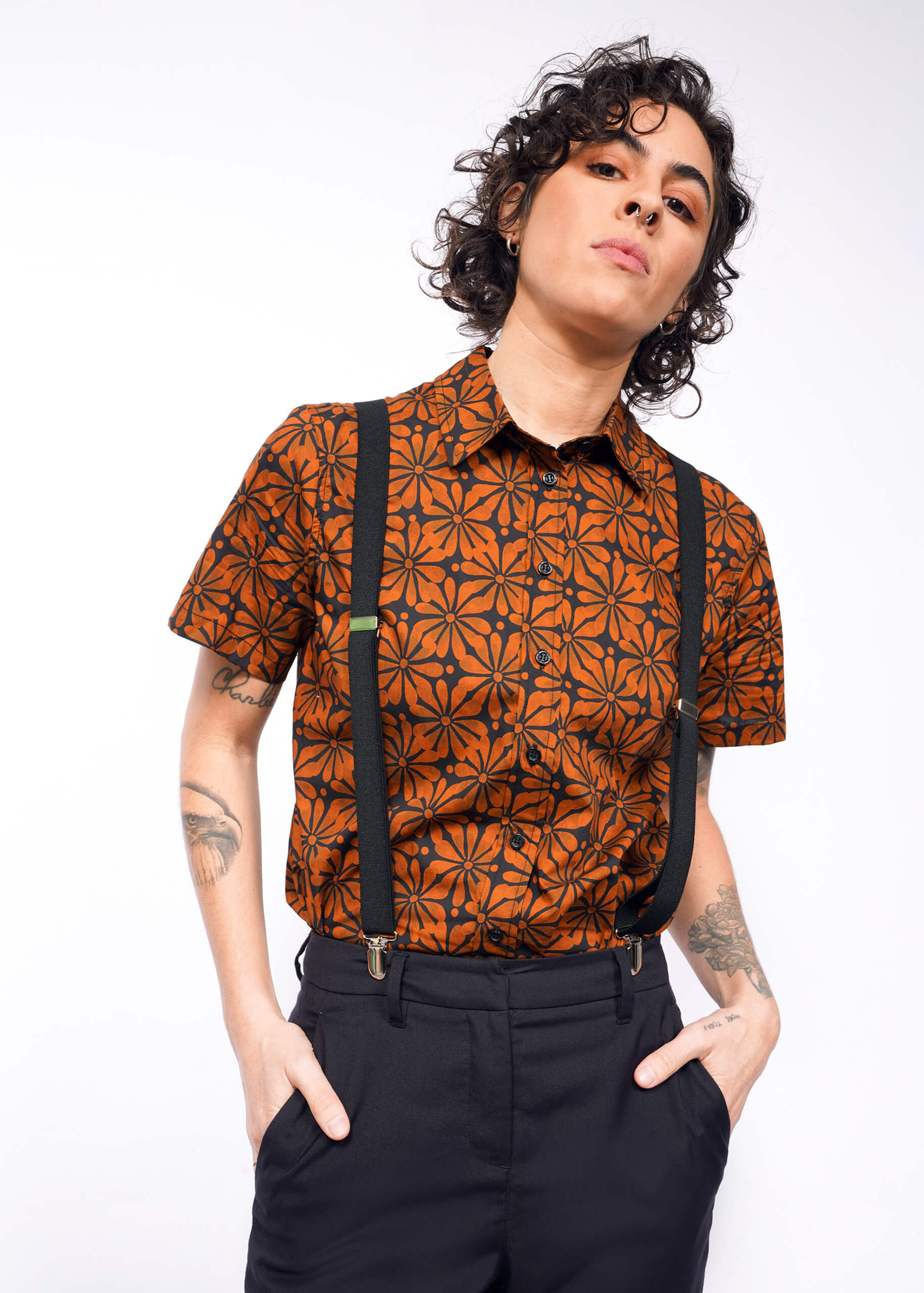 A person with curly hair is seen wearing The Essential Button Up, featuring an orange pattern and black suspenders. Tailored for a perfect fit, they stand with hands in pockets, revealing forearm tattoos against a plain white backdrop. The breathable cotton fabric enhances comfort.