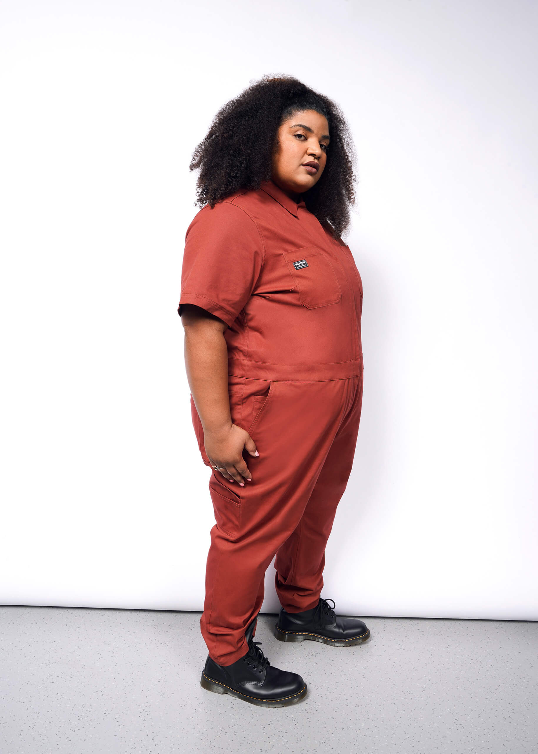 A person with curly hair stands sideways, wearing The Essential Coverall in rust stretch cotton and black boots. The relaxed fit complements their look as they pose against a plain white background, gazing at the camera with a neutral expression.