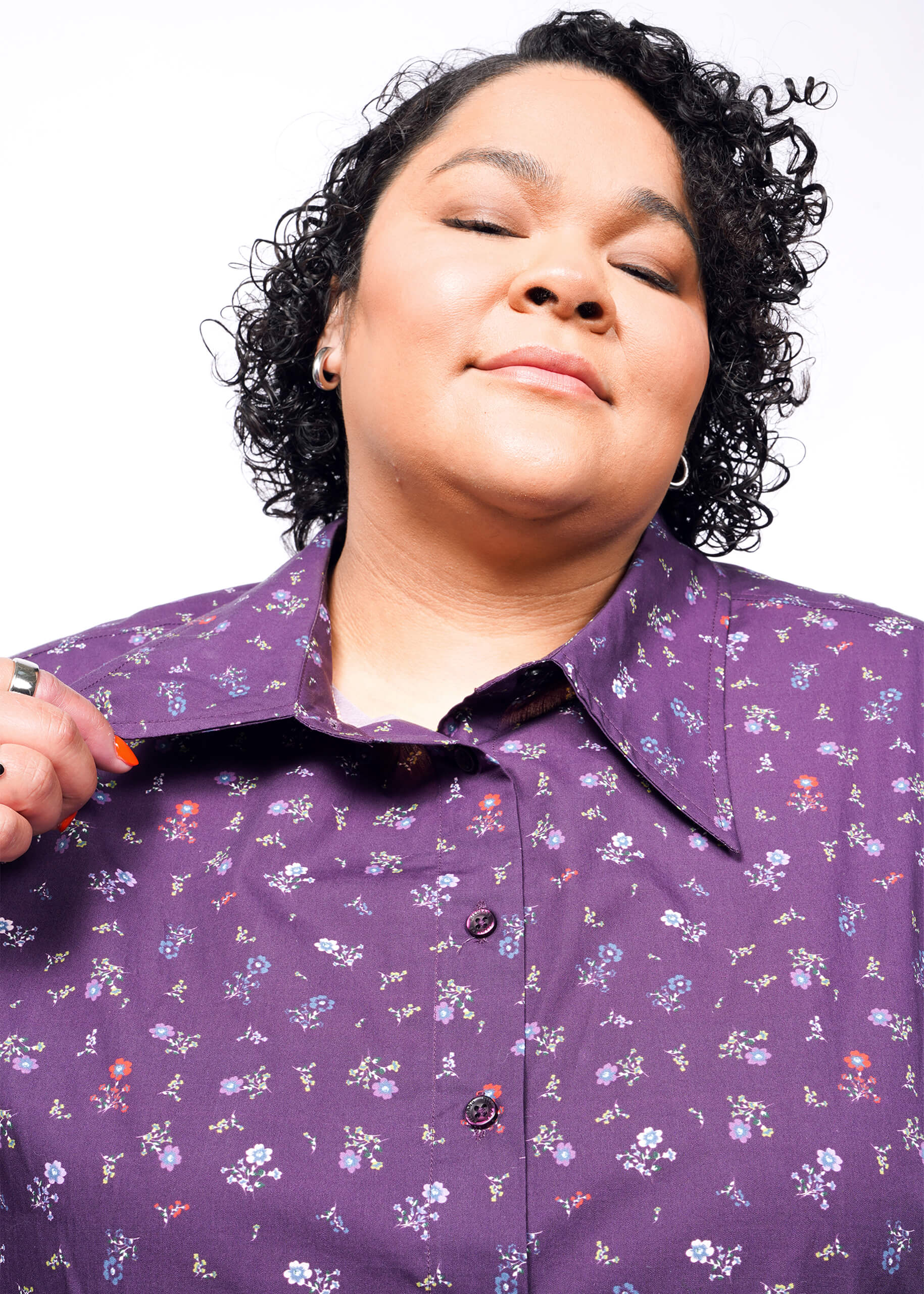 A confident individual with curly hair wears an oversized, purple floral cotton Essential Cropped Button Up. They slightly tilt their head back, holding the collar featuring distinctive Wildfang branded buttons, against a plain white background.