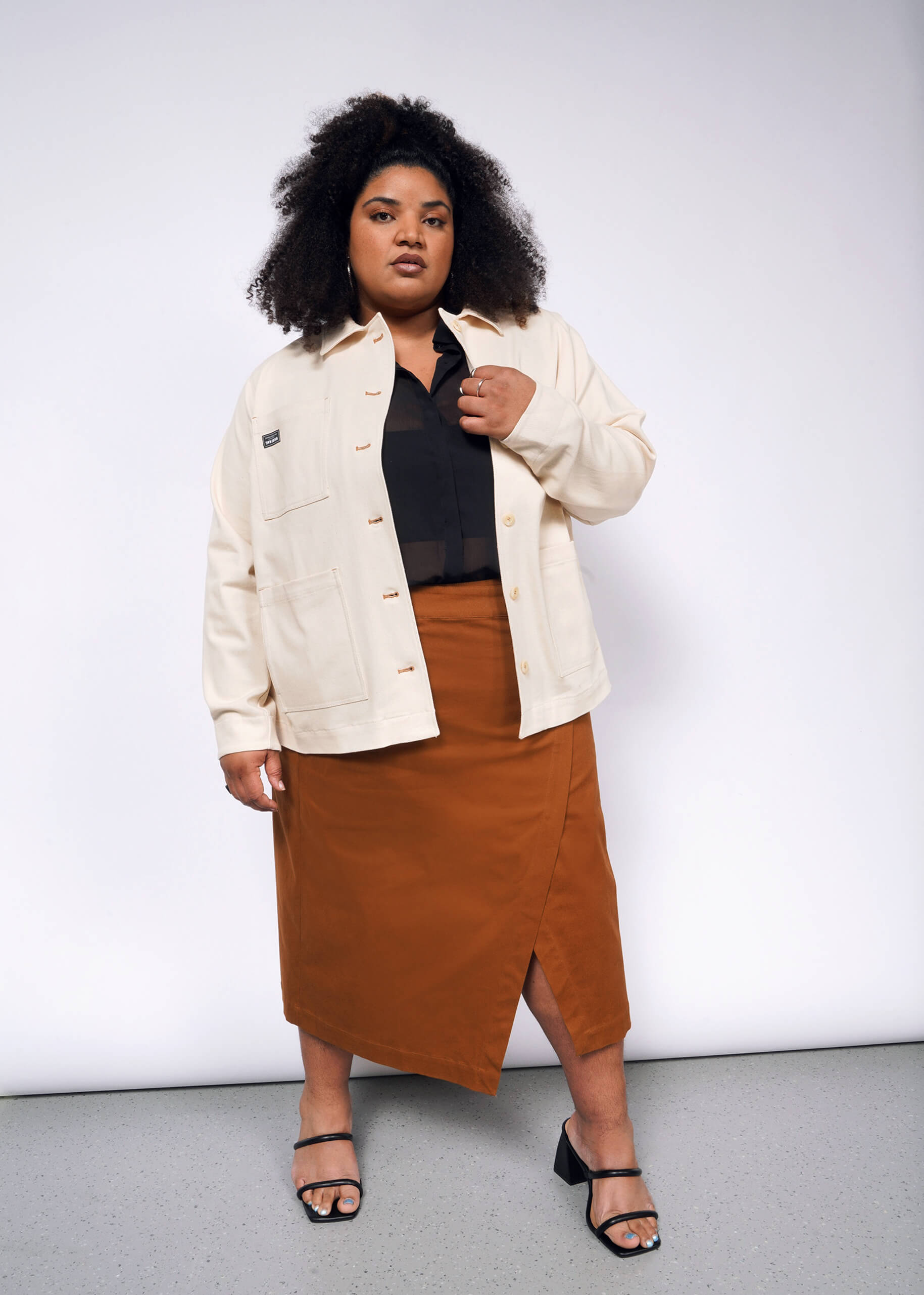 A person with curly hair stands confidently against a white background, wearing The Essential Denim Utility Jacket over a black shirt and rust-colored skirt. They sport black sandals, with one hand in the jackets deep pocket and the other touching the collar.