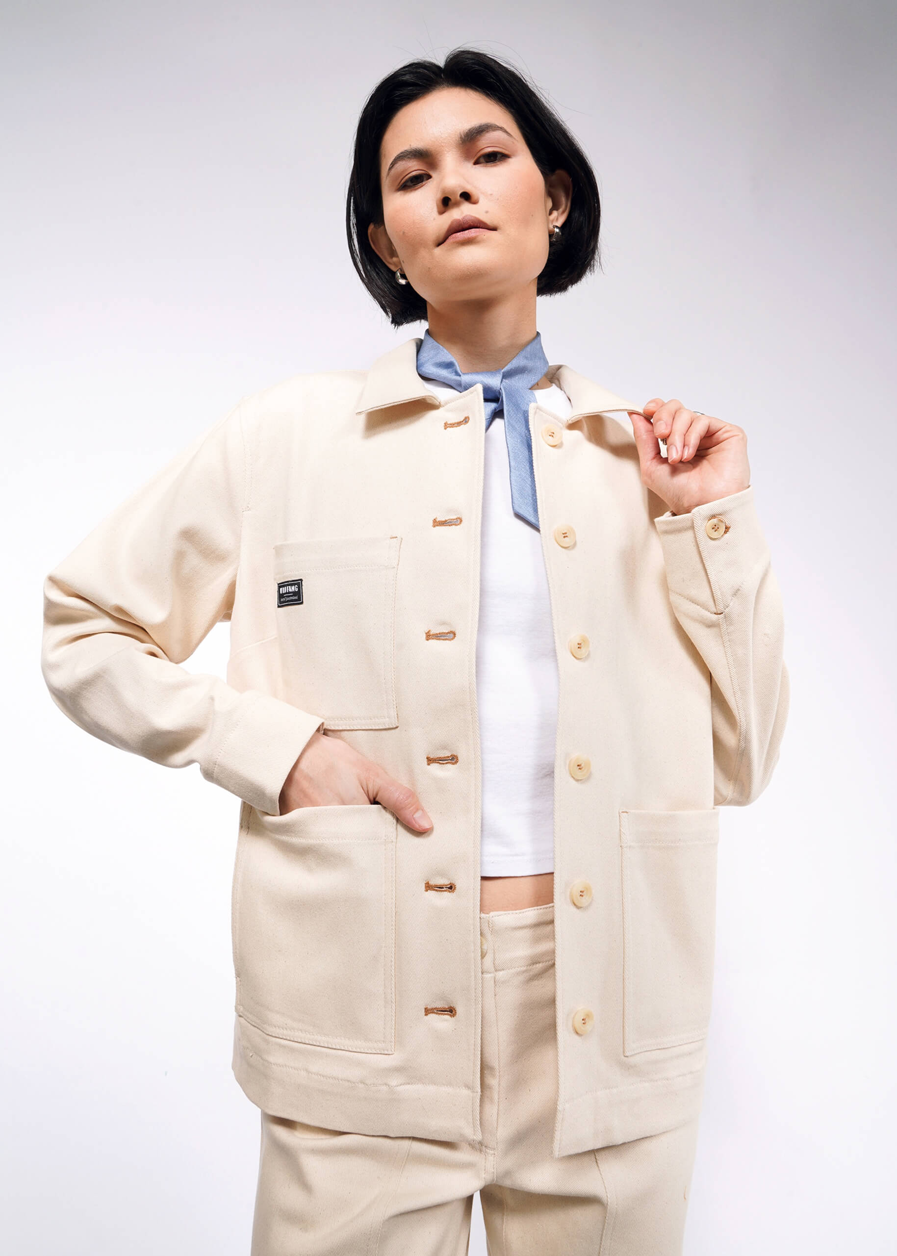 A person with short black hair wears The Essential Denim Utility Jacket in beige, featuring deep pockets over a white shirt. They pose confidently against a plain white background.