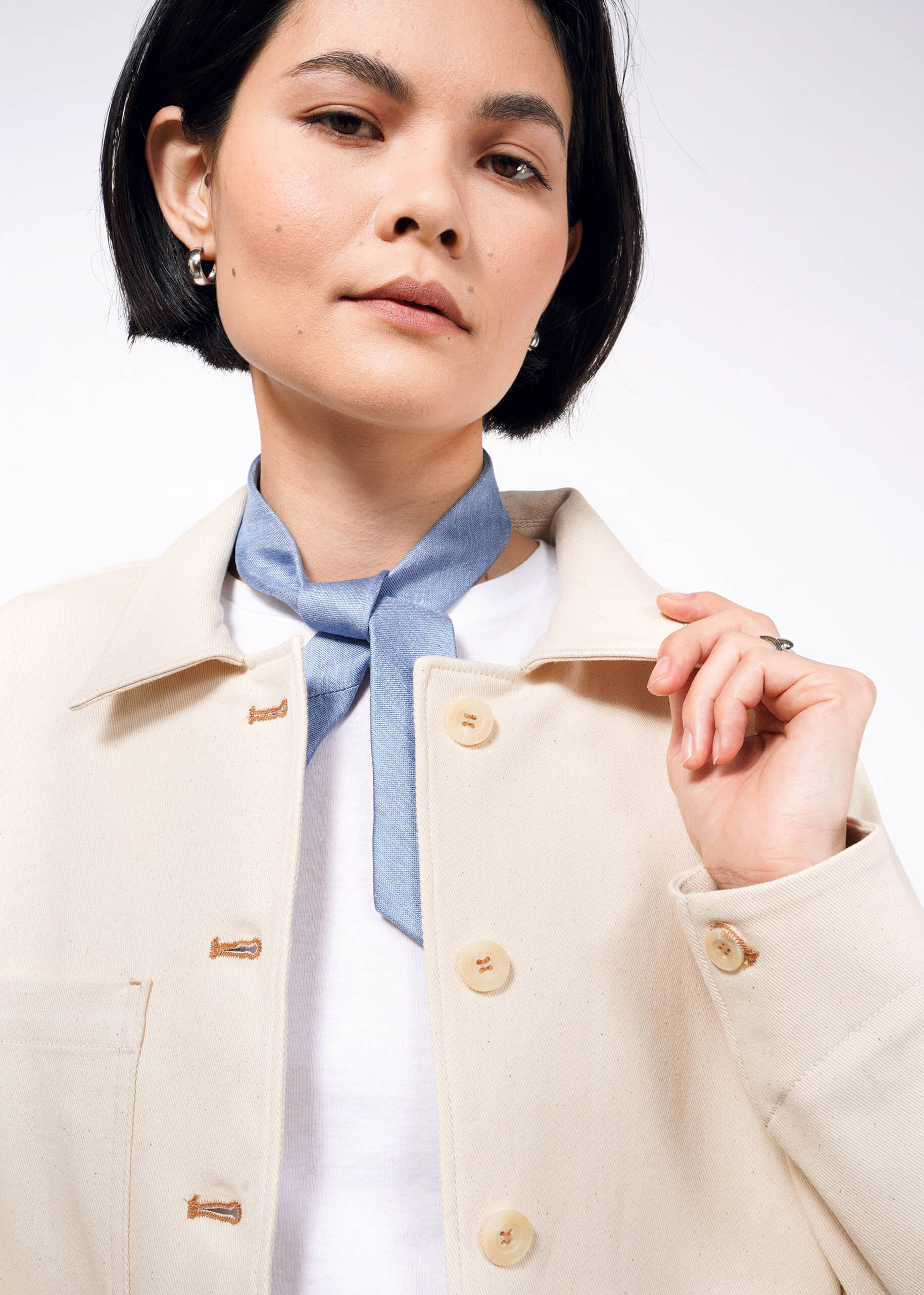 A woman with short black hair wears The Essential Denim Utility Jacket, featuring large buttons and deep pockets. She accessorizes with a blue neck scarf and small hoop earrings, holding the collar and looking slightly upward against a white background.