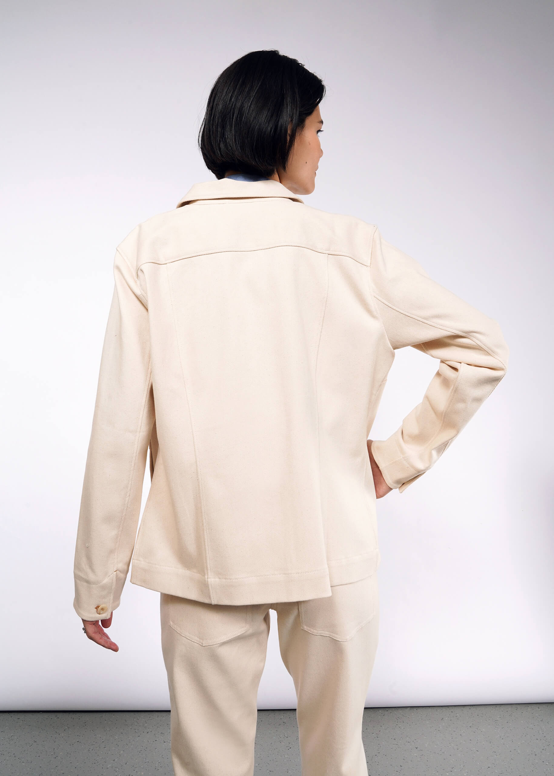 A person with short dark hair stands back to the camera wearing The Essential Denim Utility Jacket and matching pants in light beige. The outfit offers stretch comfort for all-day wear, highlighted against a plain light gray background, enhancing the sleek design of the ensemble.