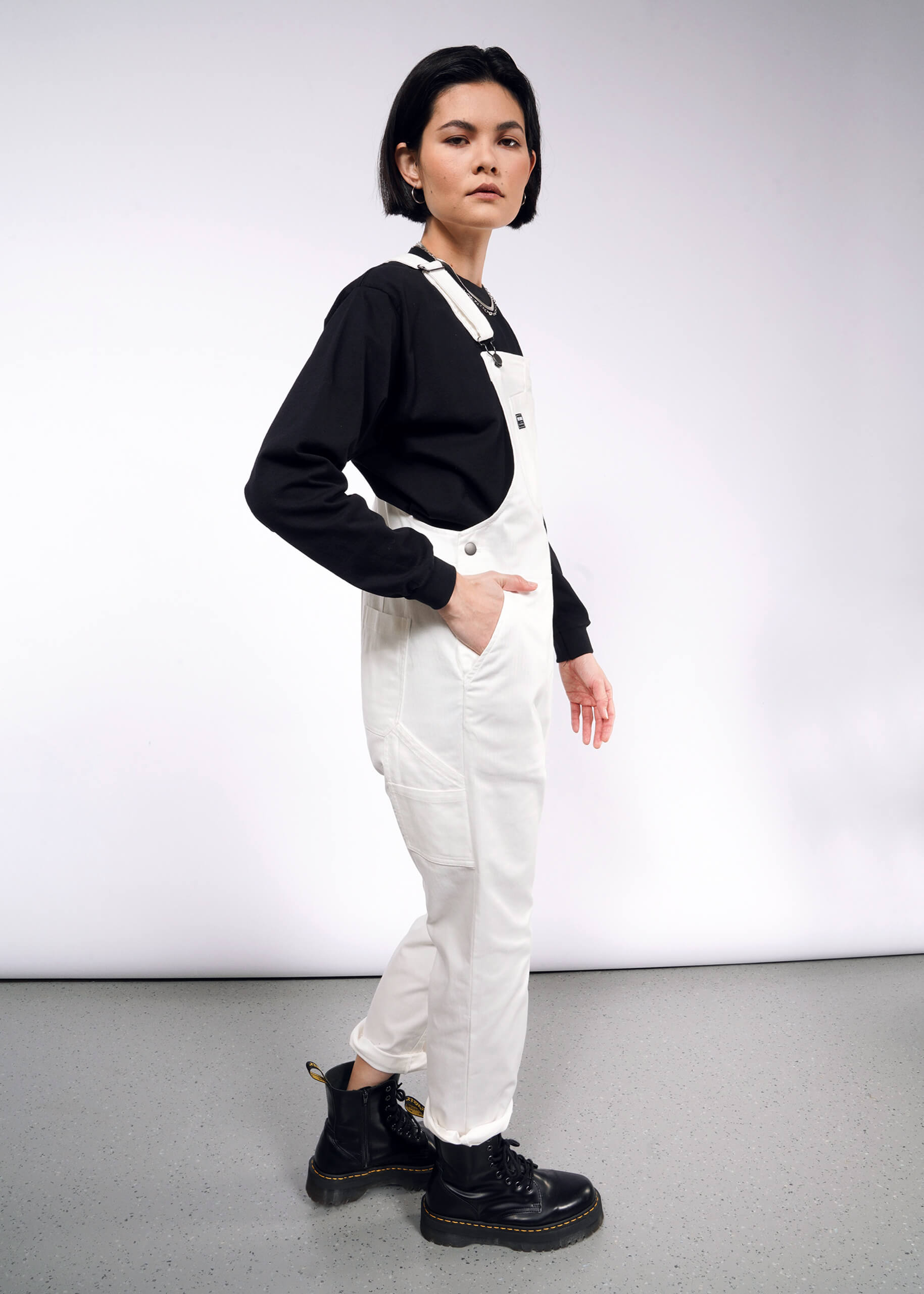 In a studio with a plain white background, an individual with short dark hair models The Essential Herringbone Overall, made of cotton. They pair it with a black long-sleeve shirt and boots, showcasing its adjustable straps and deep pocket as they stand confidently.