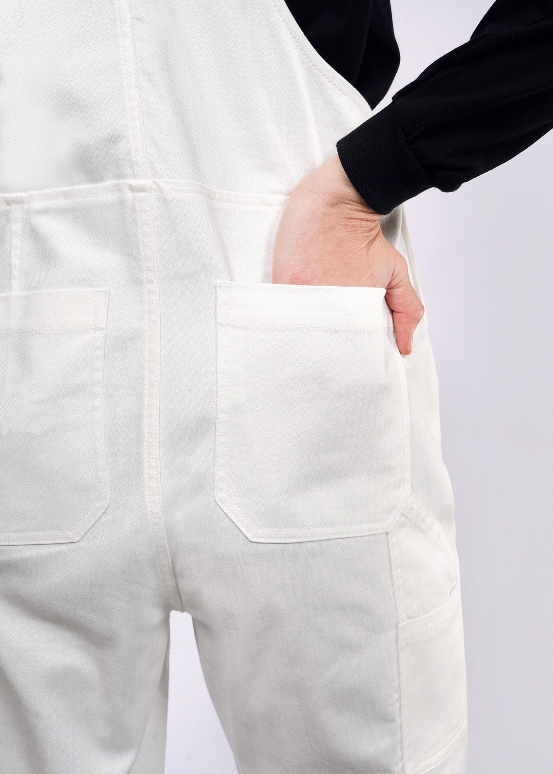 A person wearing The Essential Herringbone Overall in white cotton with a black top has their hand in the deep back pocket, showcasing the fabrics texture and stitching against a plain background, highlighting the lower back.