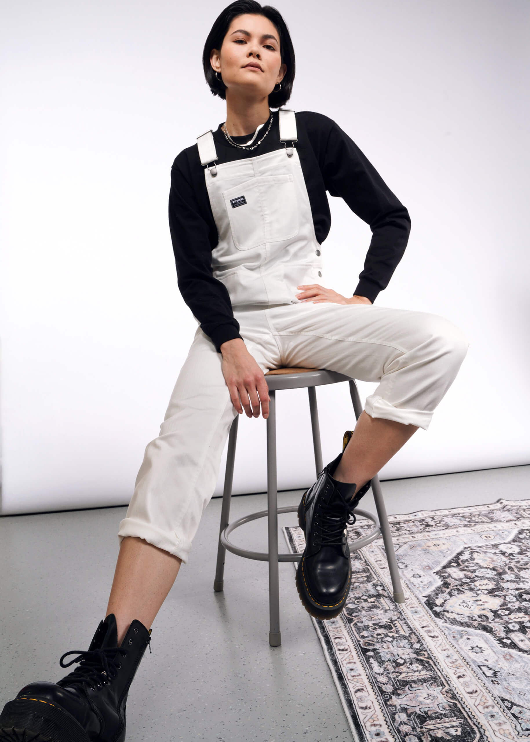 A person confidently sits on a stool wearing the Essential Herringbone Overall with adjustable straps, a black long-sleeve shirt, and sturdy black boots. Their stylish pose on a patterned rug beautifully contrasts with the simple white and gray backdrop.