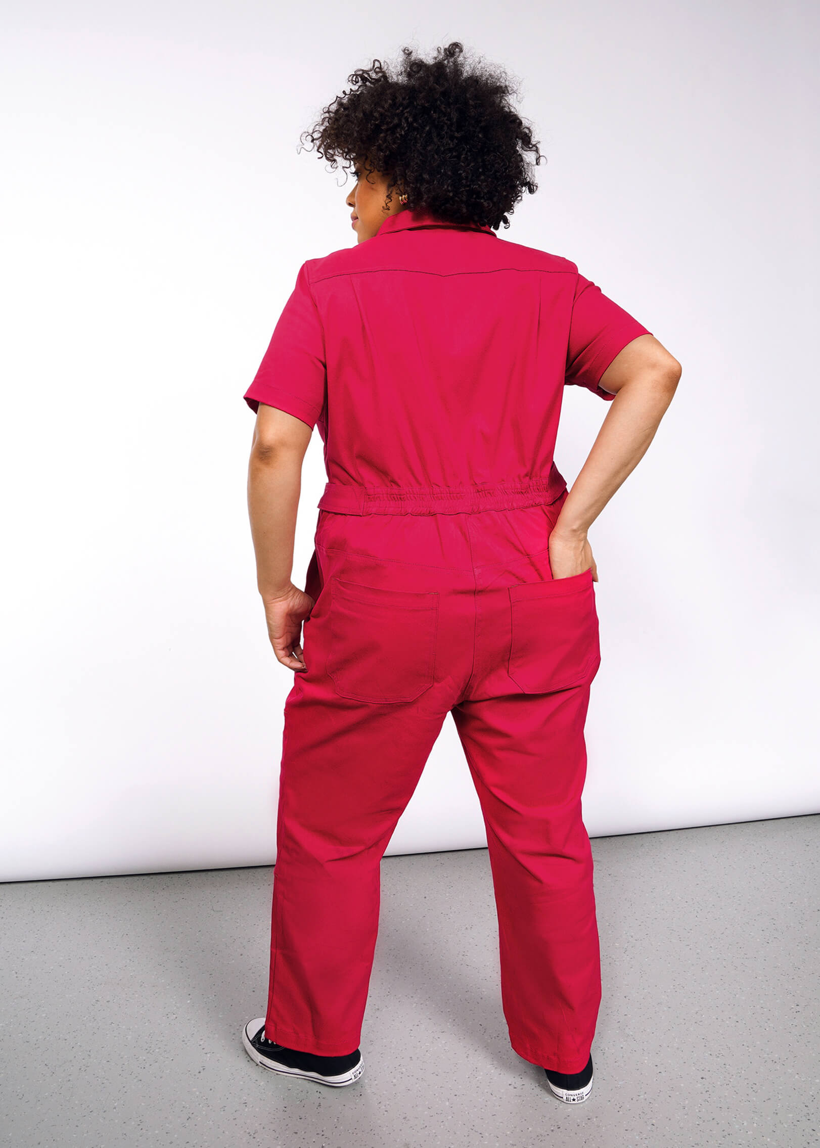 On a plain white background, a curly-haired individual wears The Essential High Waisted Coverall in bright pink paired with black sneakers, its adjustable waist and high-waisted fit elegantly highlighting their silhouette as they confidently face away on the light gray floor.