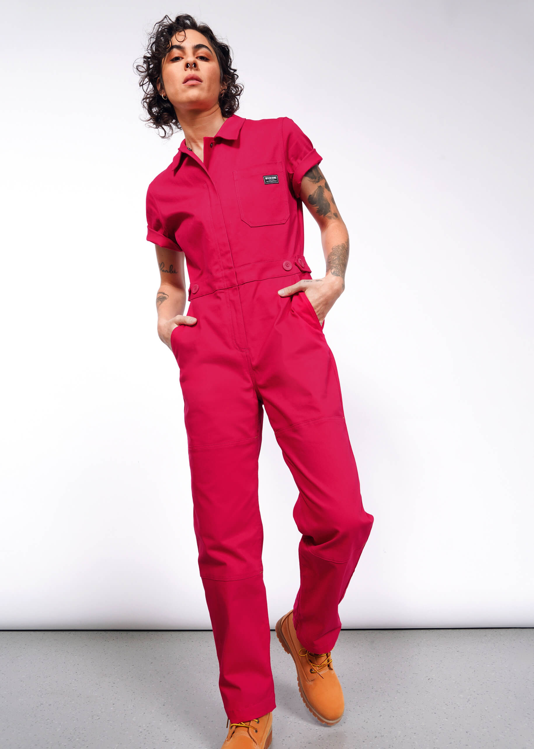 A person with curly hair confidently stands in The Essential High Waisted Coverall, a bright pink jumpsuit with an adjustable waist and deep pockets, paired with tan work boots. Against a white background, they have tattoos on their arms and hands in pockets, exuding style with a slight lean and head tilt.