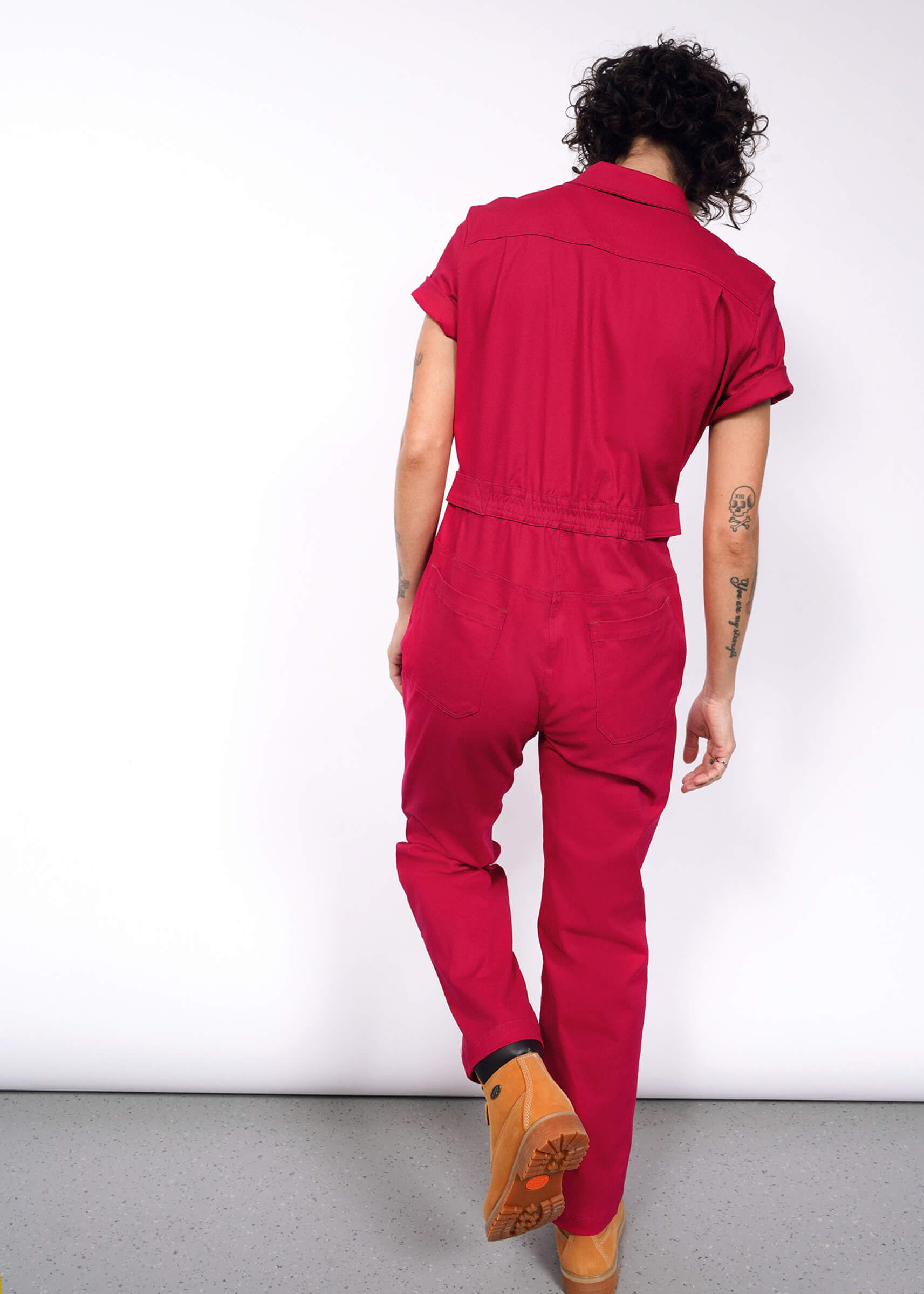 In a vibrant red Essential High Waisted Coverall with adjustable waist and deep pockets, a person with curly hair walks away from the camera. Their left arm tattoos are visible through short sleeves as they stride confidently in tan boots against a plain white background.