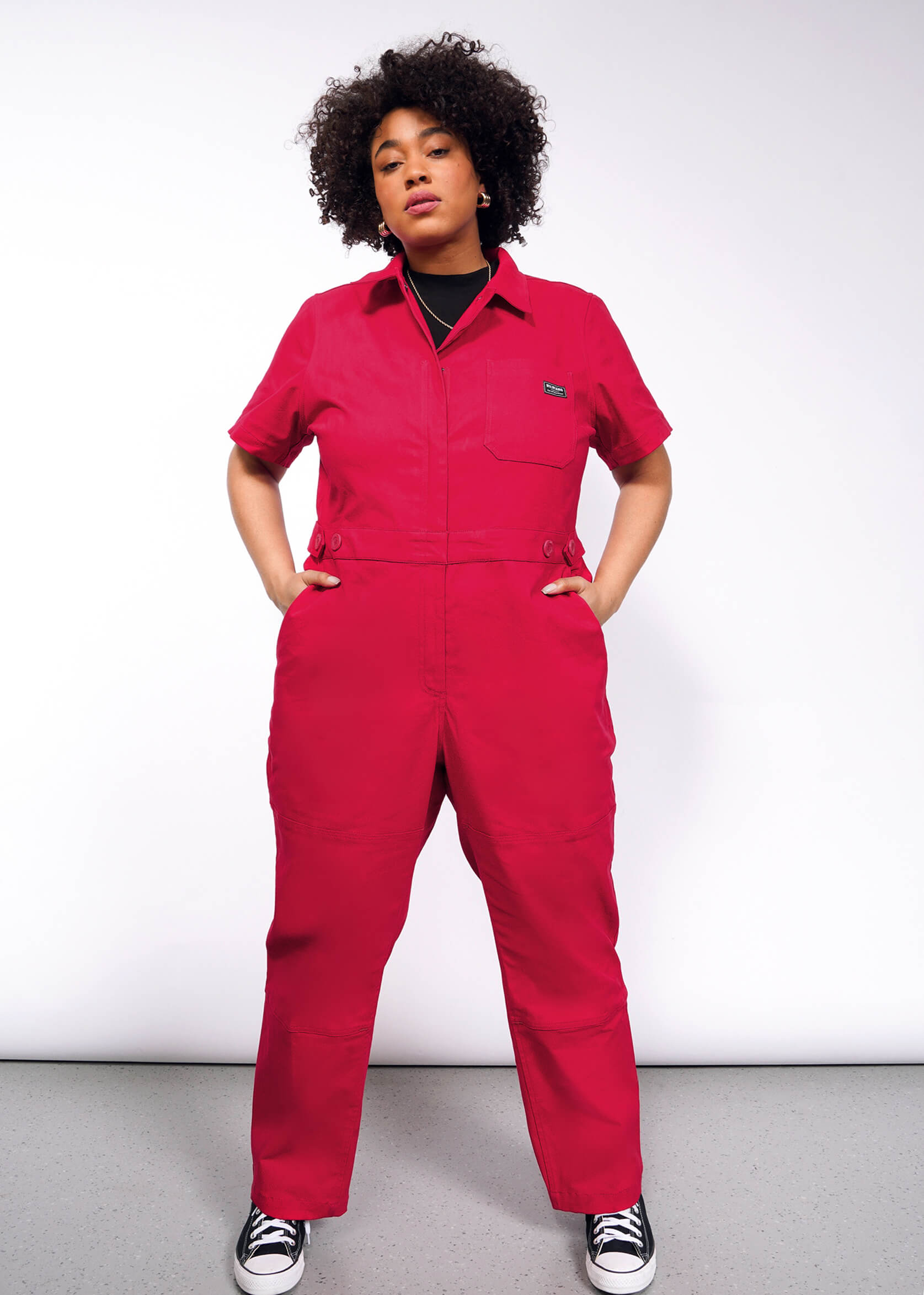 A person poses confidently against a plain backdrop, styled in The Essential High Waisted Coverall—a bright red jumpsuit with an adjustable waist—paired with black-and-white sneakers. Their hands rest in deep pockets, and they sport curly hair.