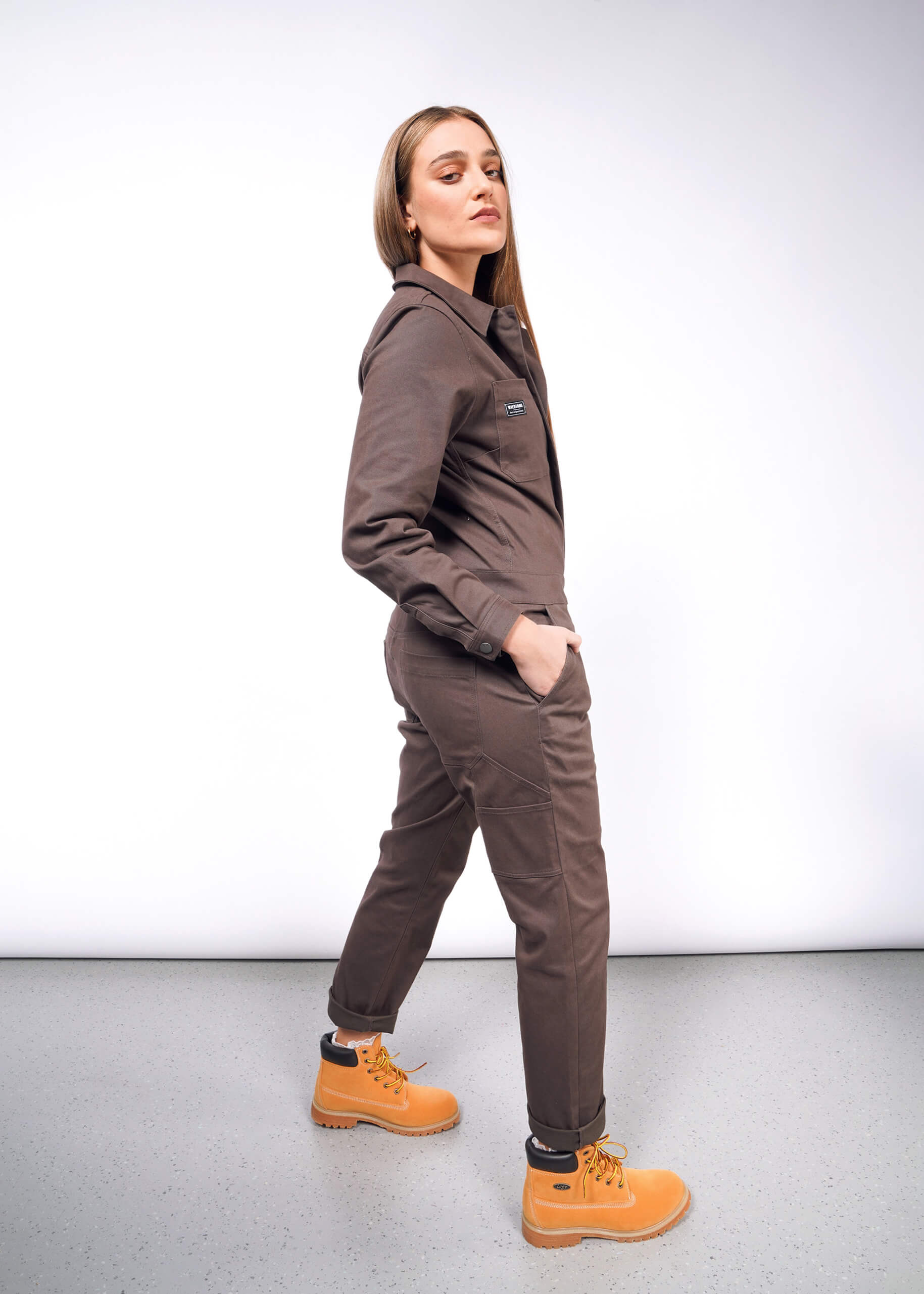 Against a white backdrop, someone sports The Essential Long Sleeve Coverall in oversized brushed stretch cotton, along with sturdy yellow work boots. With hands casually in pockets, they exude confidence while gazing sideways.