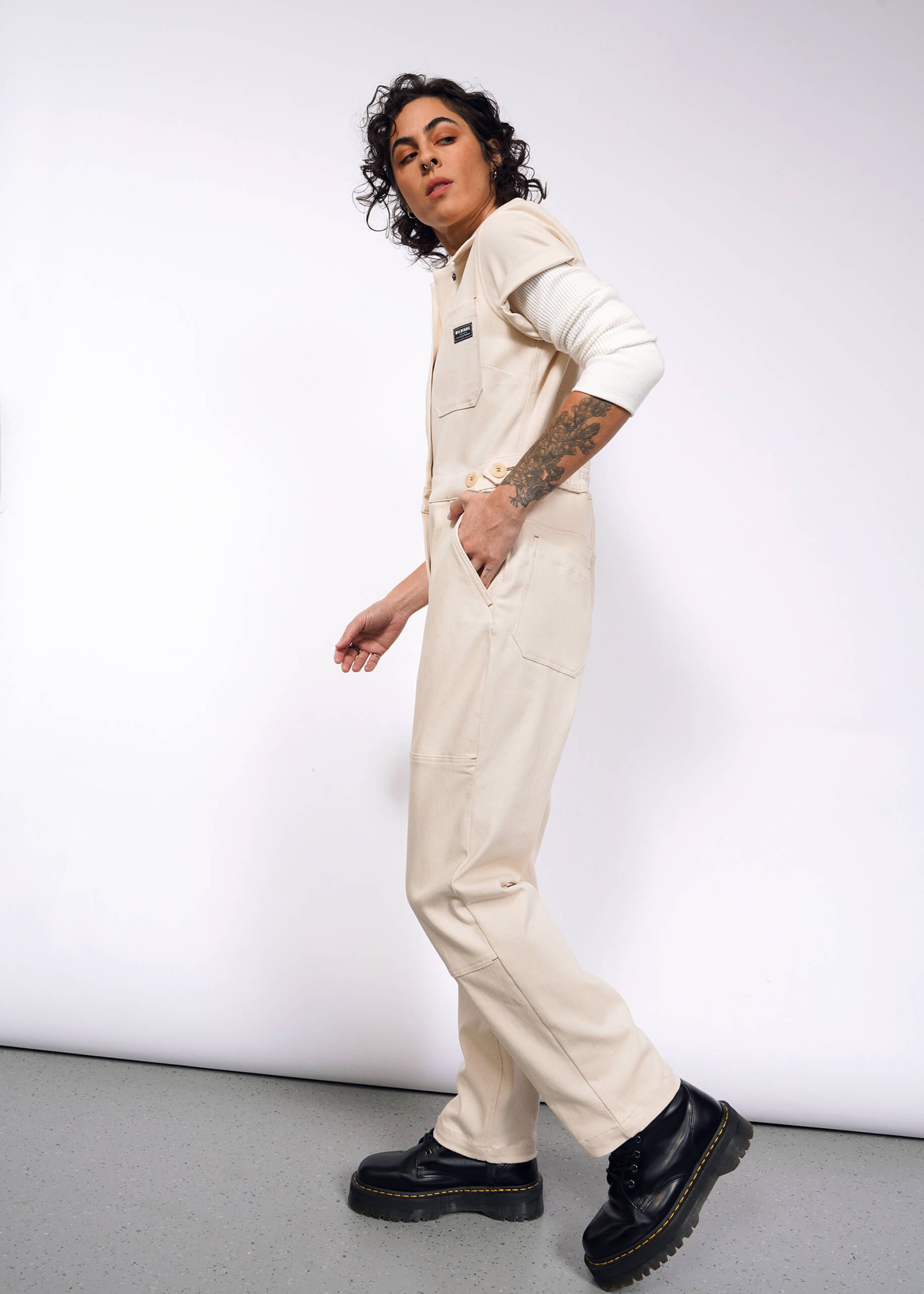 A person with curly hair models The Essential Denim High Waisted Coverall, featuring deep pockets and an undyed finish. Tattoos adorn their left arm as they tuck their right hand casually. They wear black shoes while appearing in motion against a plain backdrop.