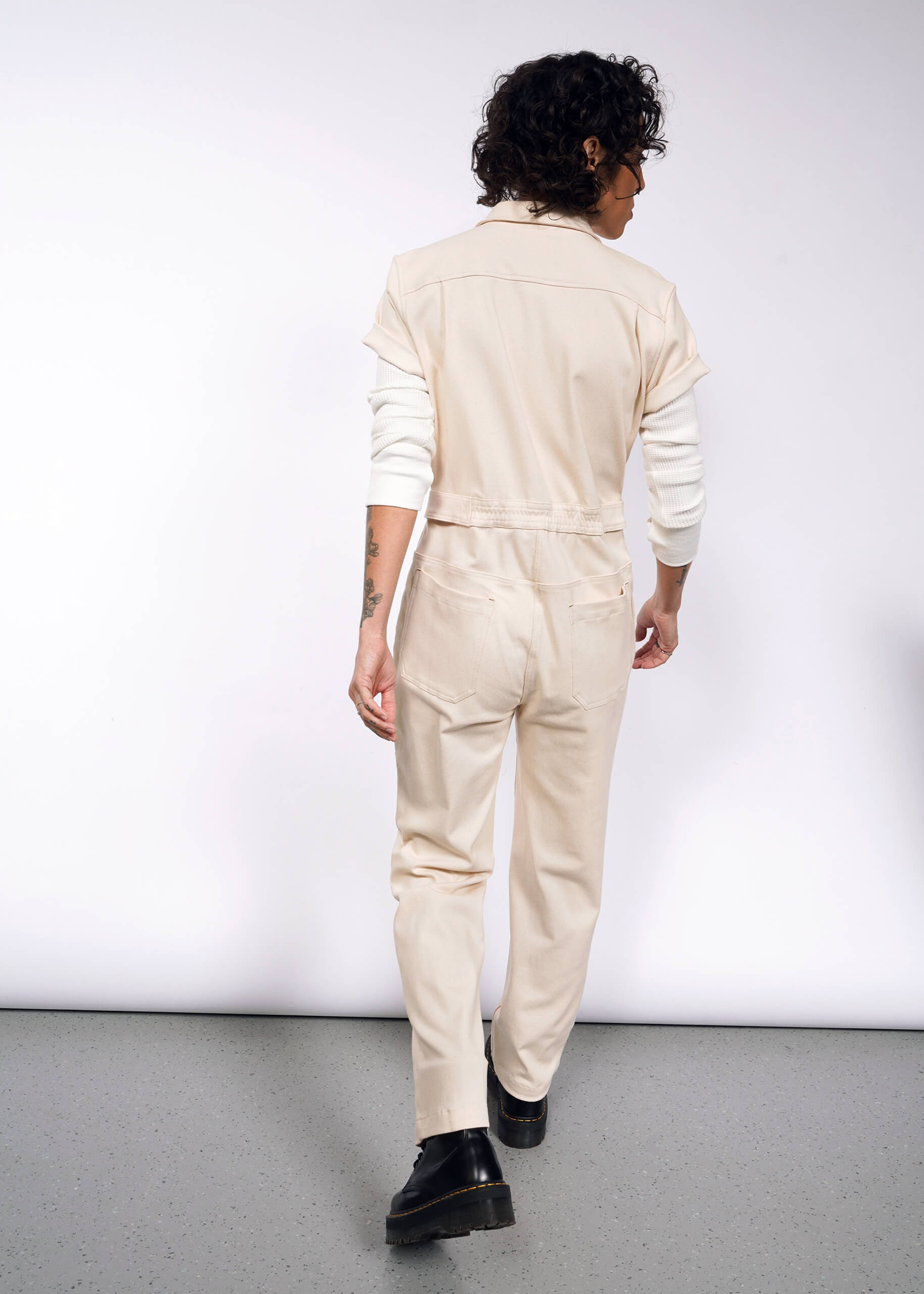 Facing away, a person with curly hair wears The Essential Denim High Waisted Coverall in cream over a long-sleeve white shirt, displaying tattoos on their arm. They walk on a speckled floor against a white backdrop, completing the look with black shoes.