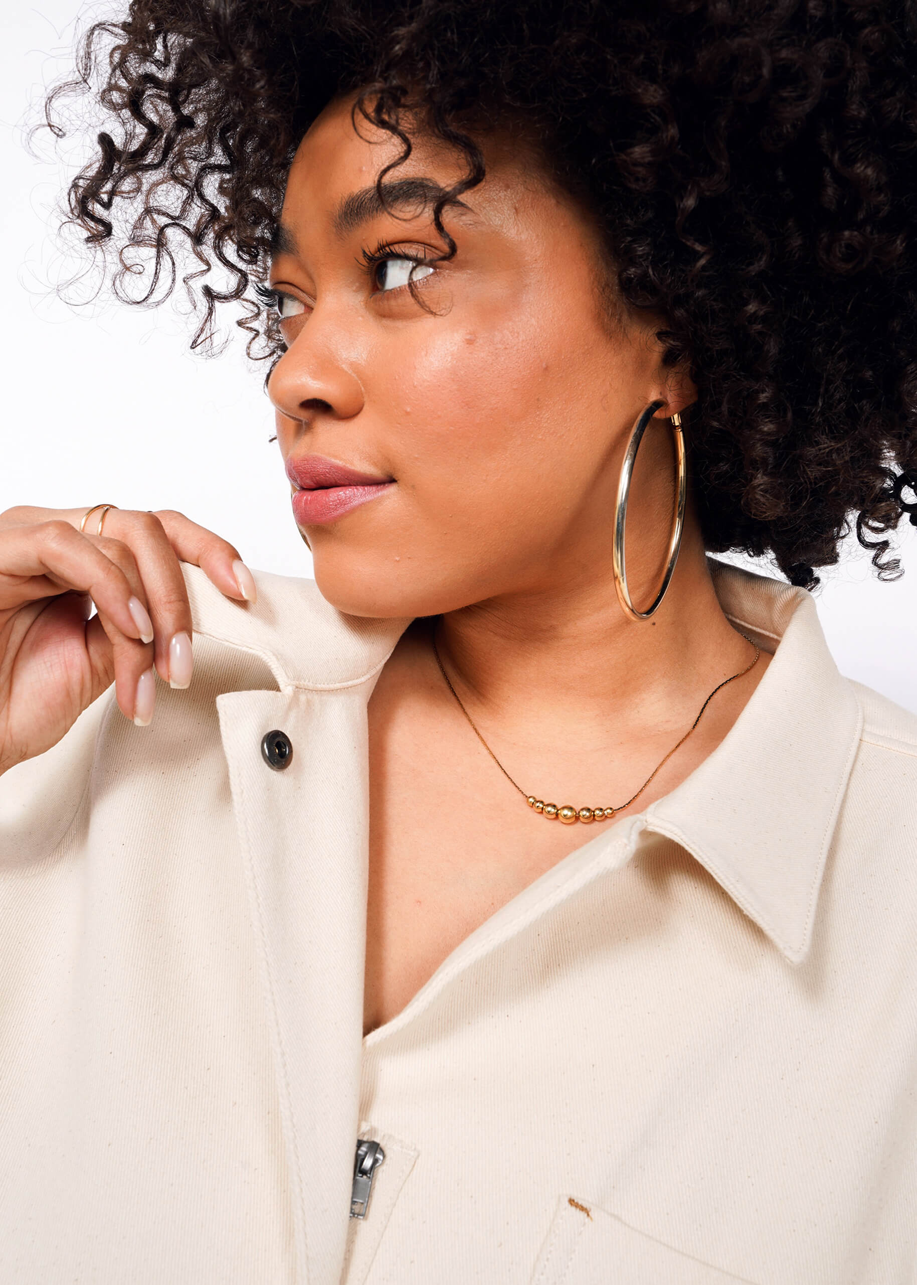 A person with curly hair, large hoop earrings, and a gold necklace is gazing to the side. Theyre holding the collar of a cream-colored jacket over The Essential Denim High Waisted Coverall. The background is plain white.