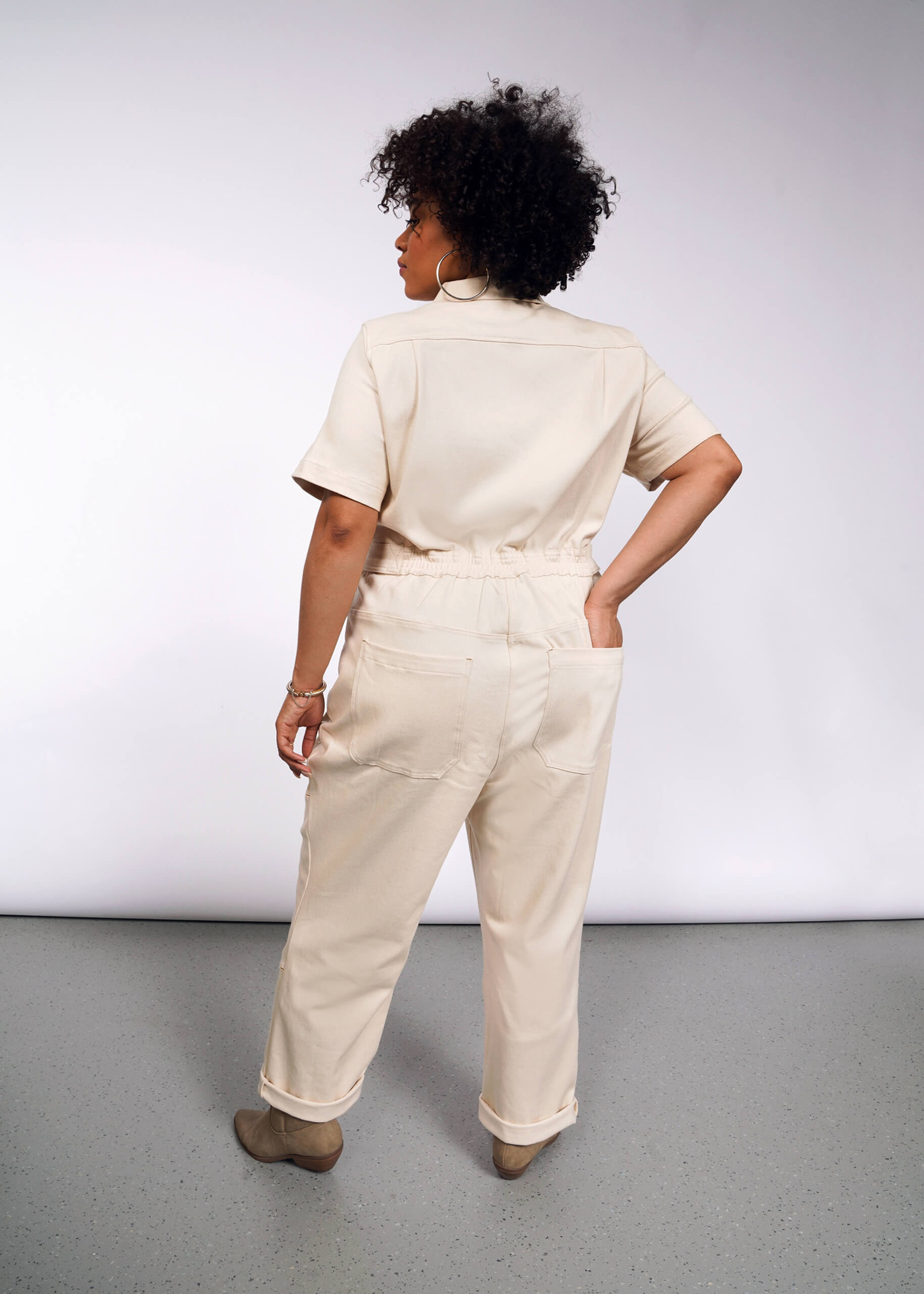 A woman with curly hair stands with her back to the camera, wearing The Essential Denim High Waisted Coverall in beige and matching shoes. She tucks her left hand into one of the deep back pockets against a gray background.