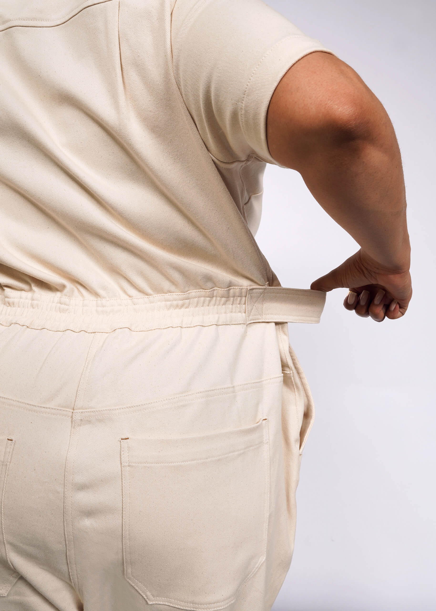 Someone wearing The Essential Denim High Waisted Coverall in cream is adjusting the waistband at the back. The short-sleeved ensemble has deep side pockets, with focus on their back and side against a plain backdrop.