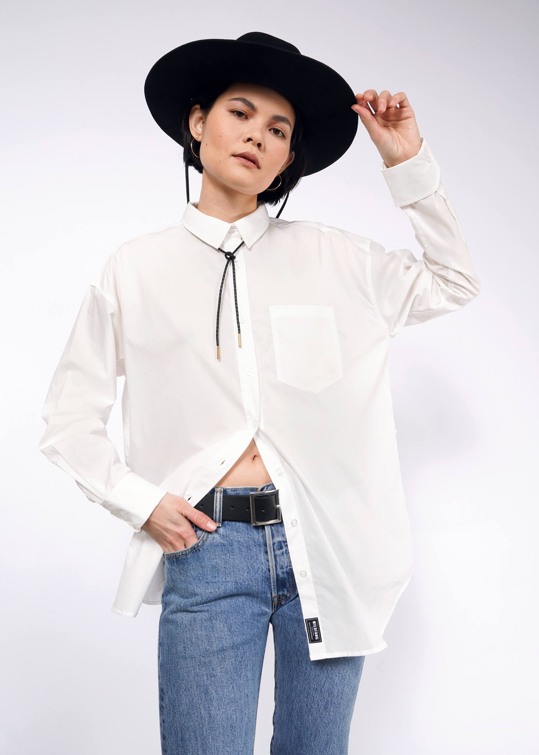 Person posing against a white background wearing The Essential Long Sleeve Oversized Wrap Button Up with French cuffs, bolo tie, blue jeans, and a wide-brimmed black hat. The shirt is partially tucked in, and they hold the brim of the hat with one hand.