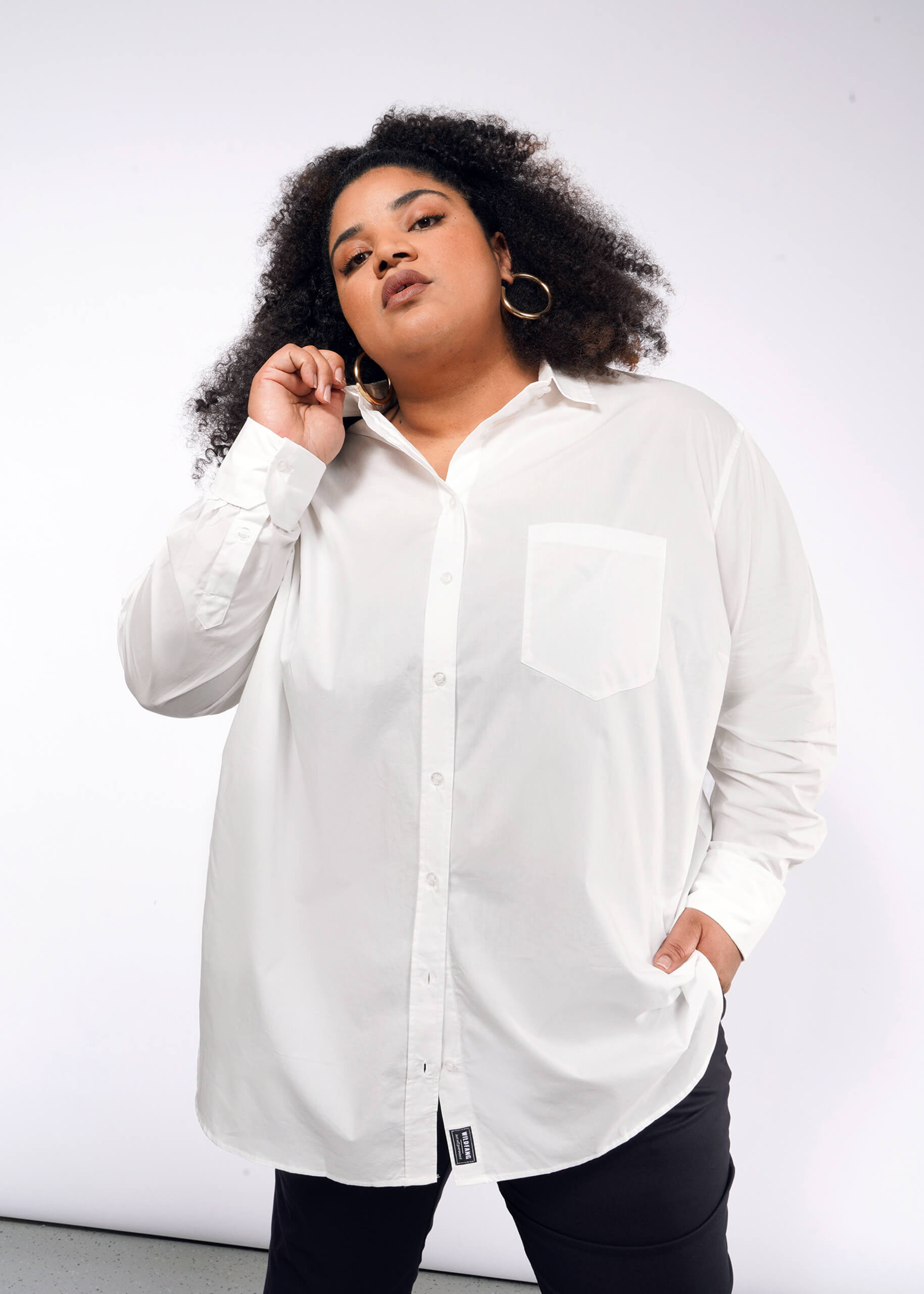 A person with curly hair and large hoop earrings poses confidently, wearing The Essential Long Sleeve Oversized Wrap Button Up shirt with French cuffs and black pants. One hand touches the collar while the other rests in a pocket against a plain white background.