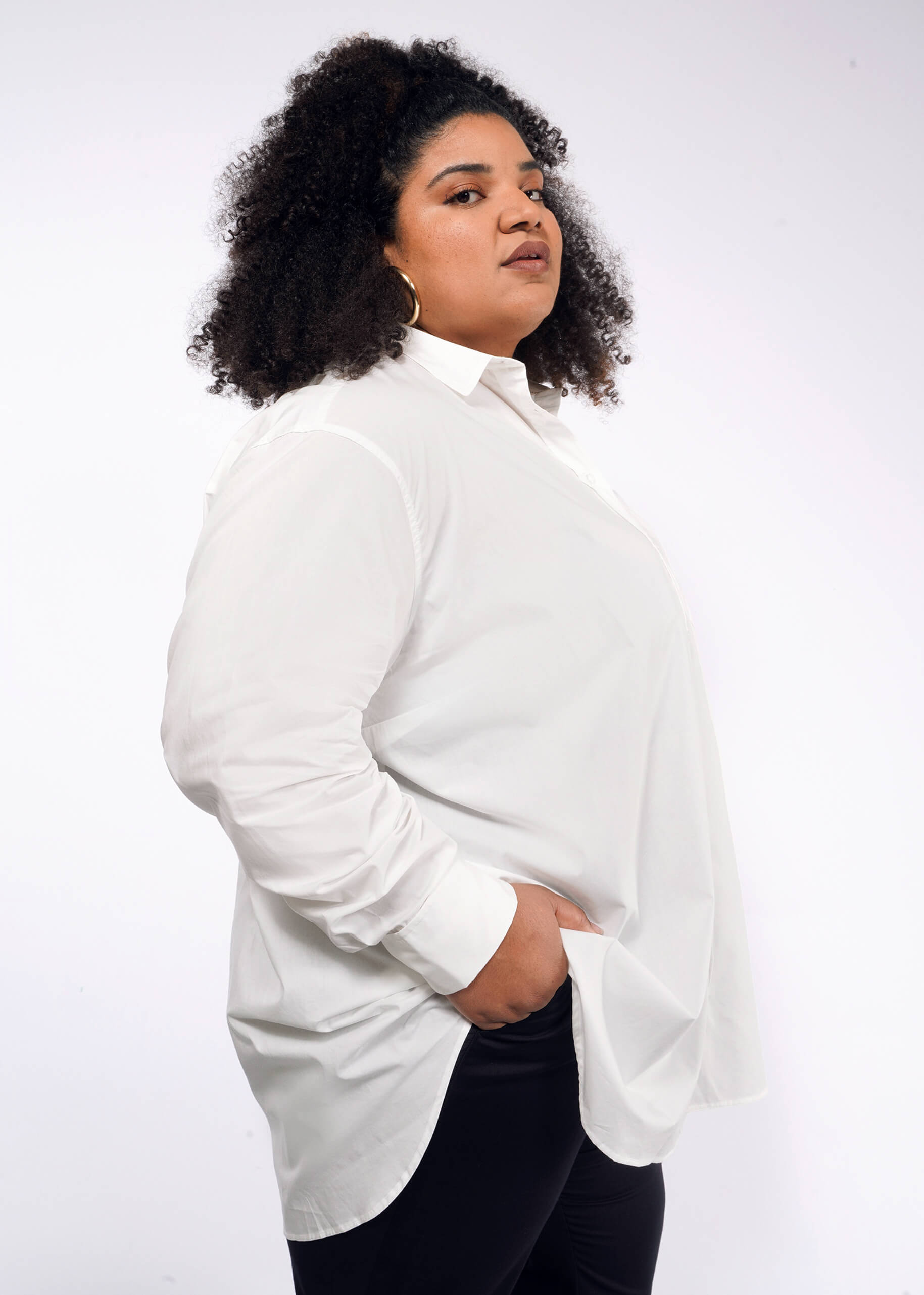 A person with curly hair confidently stands against a plain background, wearing The Essential Long Sleeve Oversized Wrap Button Up shirt and black pants, with one hand in their pocket.