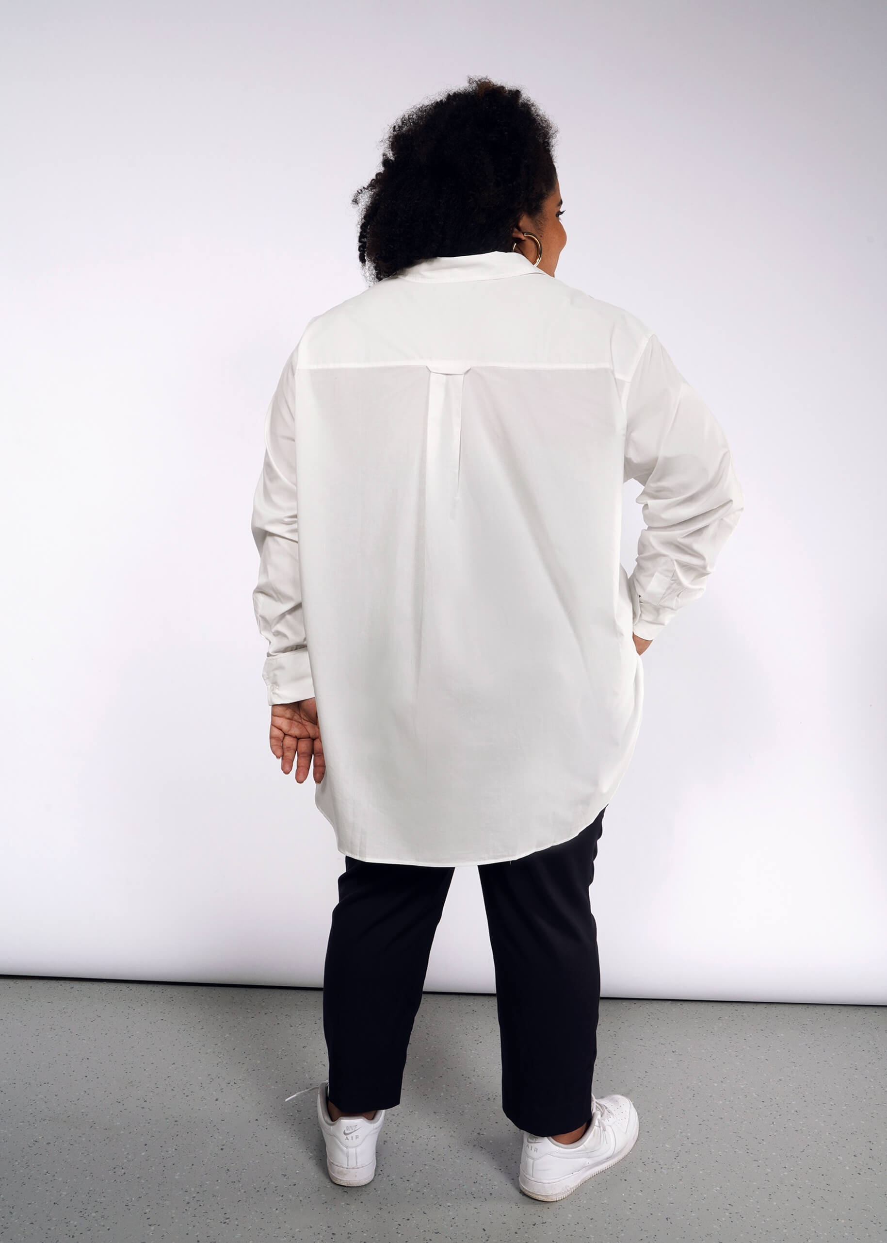 A person with curly hair, facing away from the camera, wears The Essential Long Sleeve Oversized Wrap Button Up with French cuffs, black pants, and white sneakers. A light gray wall and floor create a minimalist backdrop.