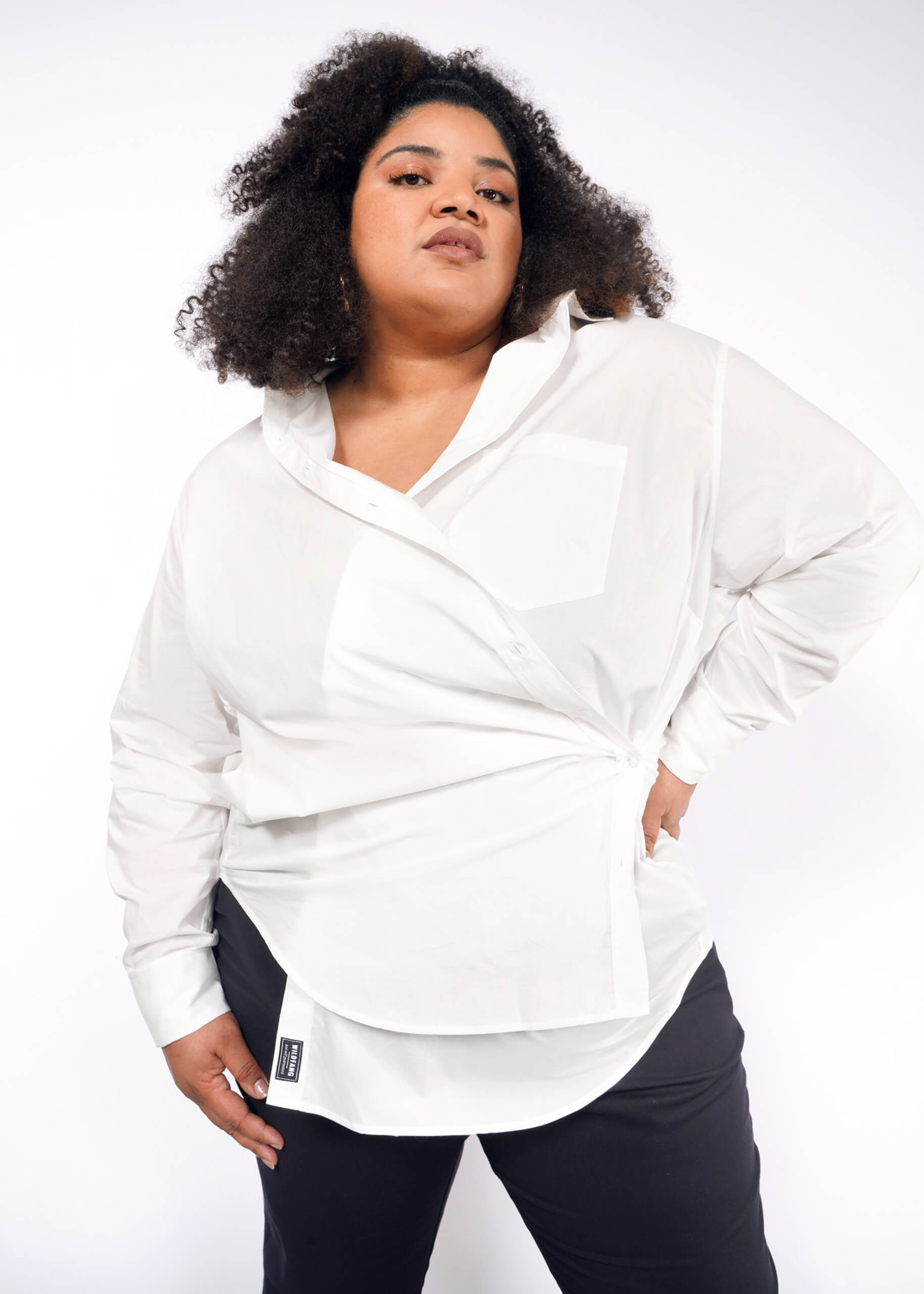 A person with voluminous curly hair confidently poses in The Essential Long Sleeve Oversized Wrap Button Up and black pants, one hand in a pocket and the other by their side against a plain white background.