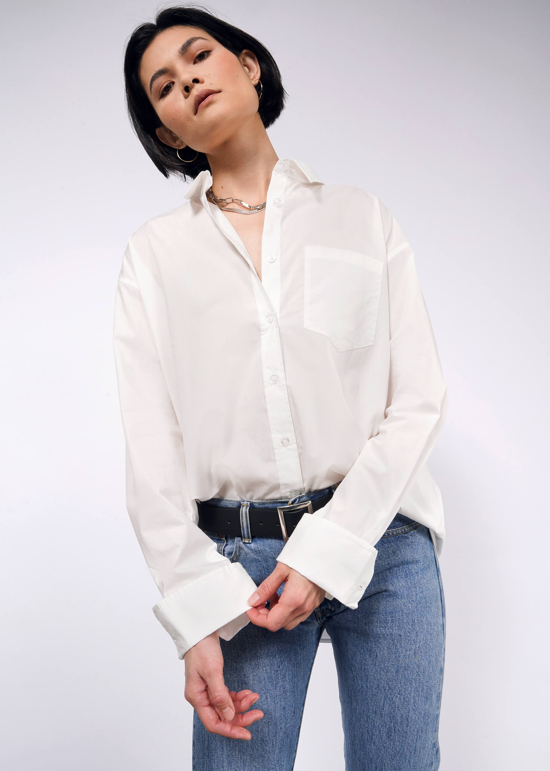 A person with short dark hair is wearing The Essential Long Sleeve Oversized Wrap Button Up in white, paired with blue jeans and a black belt. They pose against a plain background, gazing slightly downward.