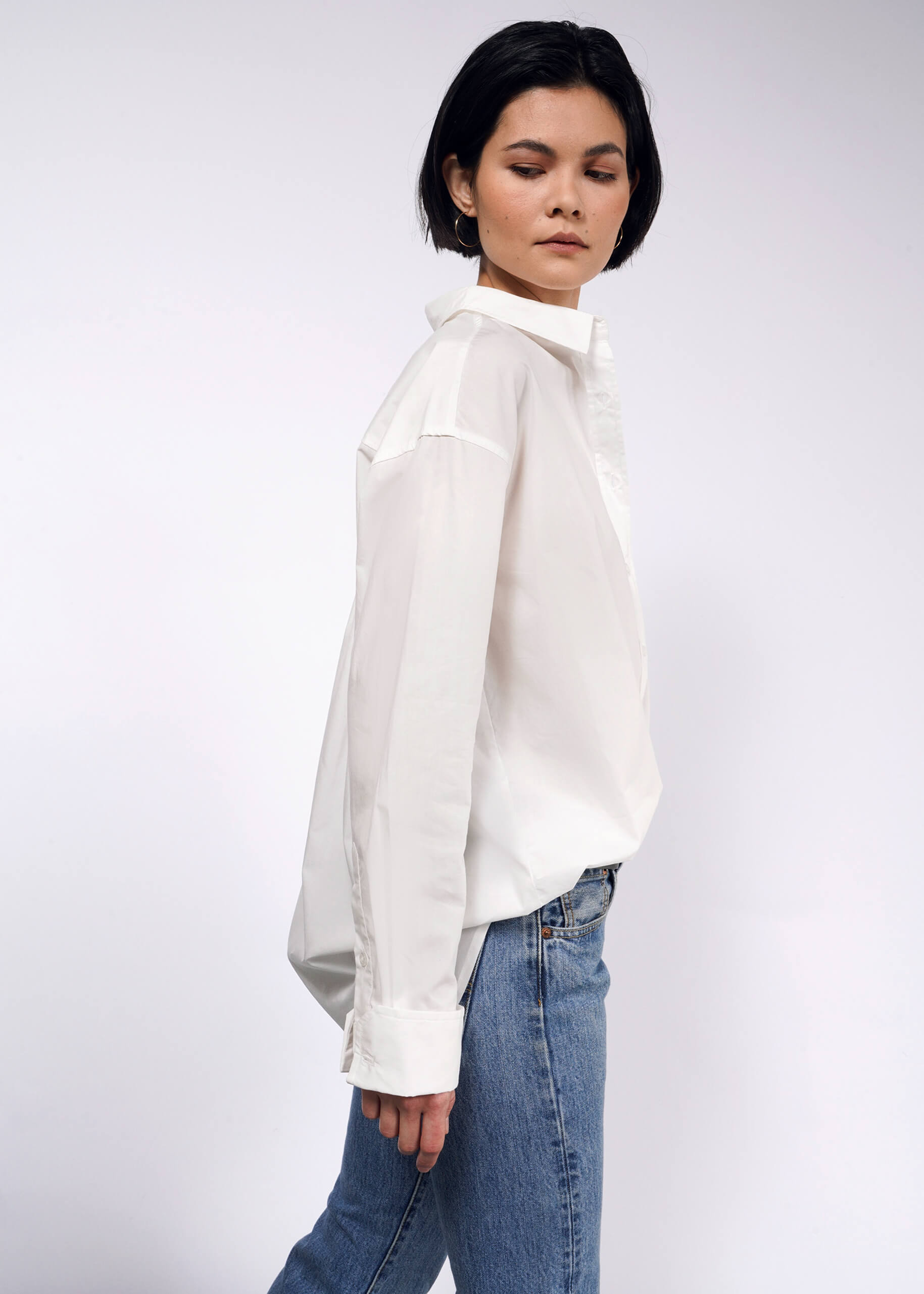 A person with short black hair is wearing The Essential Long Sleeve Oversized Wrap Button Up in white, paired with blue jeans. They stand in profile against a plain white background, looking downwards with a neutral expression.