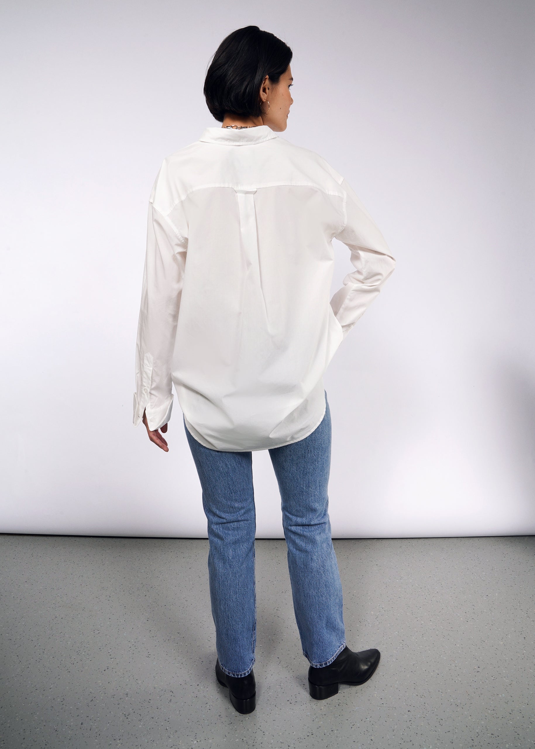 With their back to the camera, a person wears The Essential Long Sleeve Oversized Wrap Button Up, paired with blue jeans and black shoes. The background is plain and light-colored.