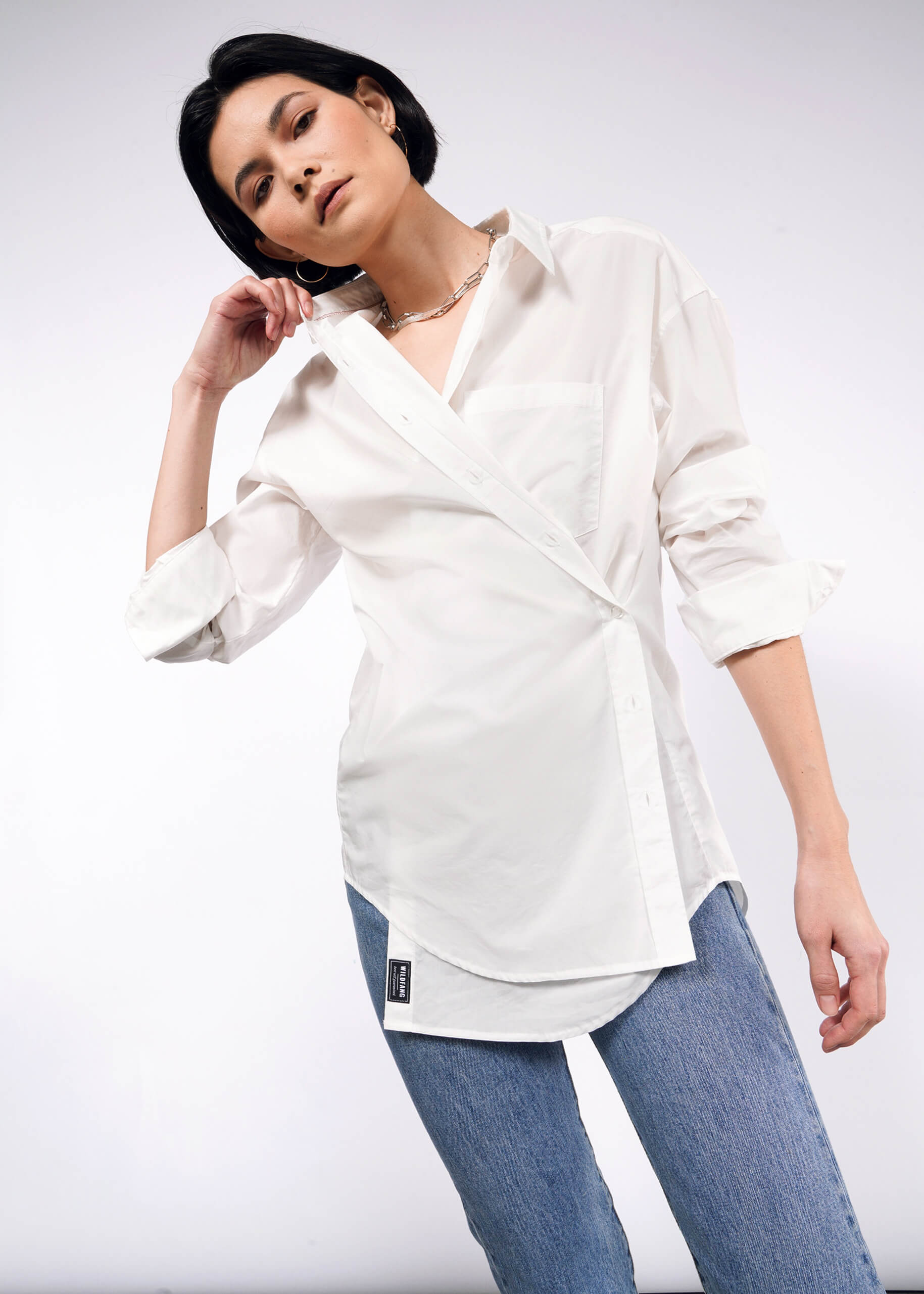 A person with short dark hair confidently poses wearing The Essential Long Sleeve Oversized Wrap Button Up featuring French cuffs and rolled-up sleeves, paired effortlessly with light blue jeans. The background is plain and light-colored.