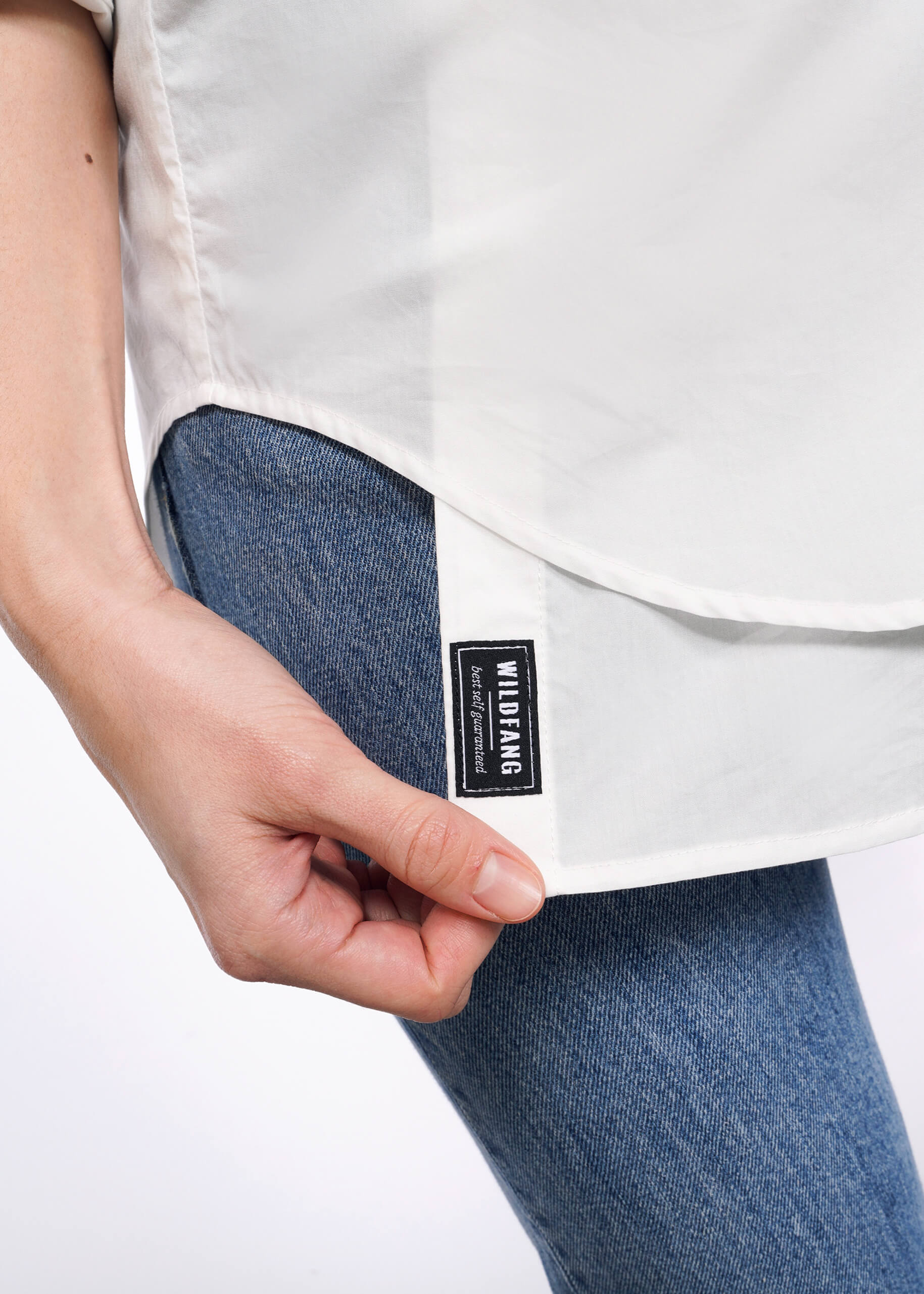 Close-up of a person holding the edge of The Essential Long Sleeve Oversized Wrap Button Up in white, featuring a black Wu-Tang Brand Limited tag. They are wearing blue jeans, with focus on the shirt and tag against a plain white background.