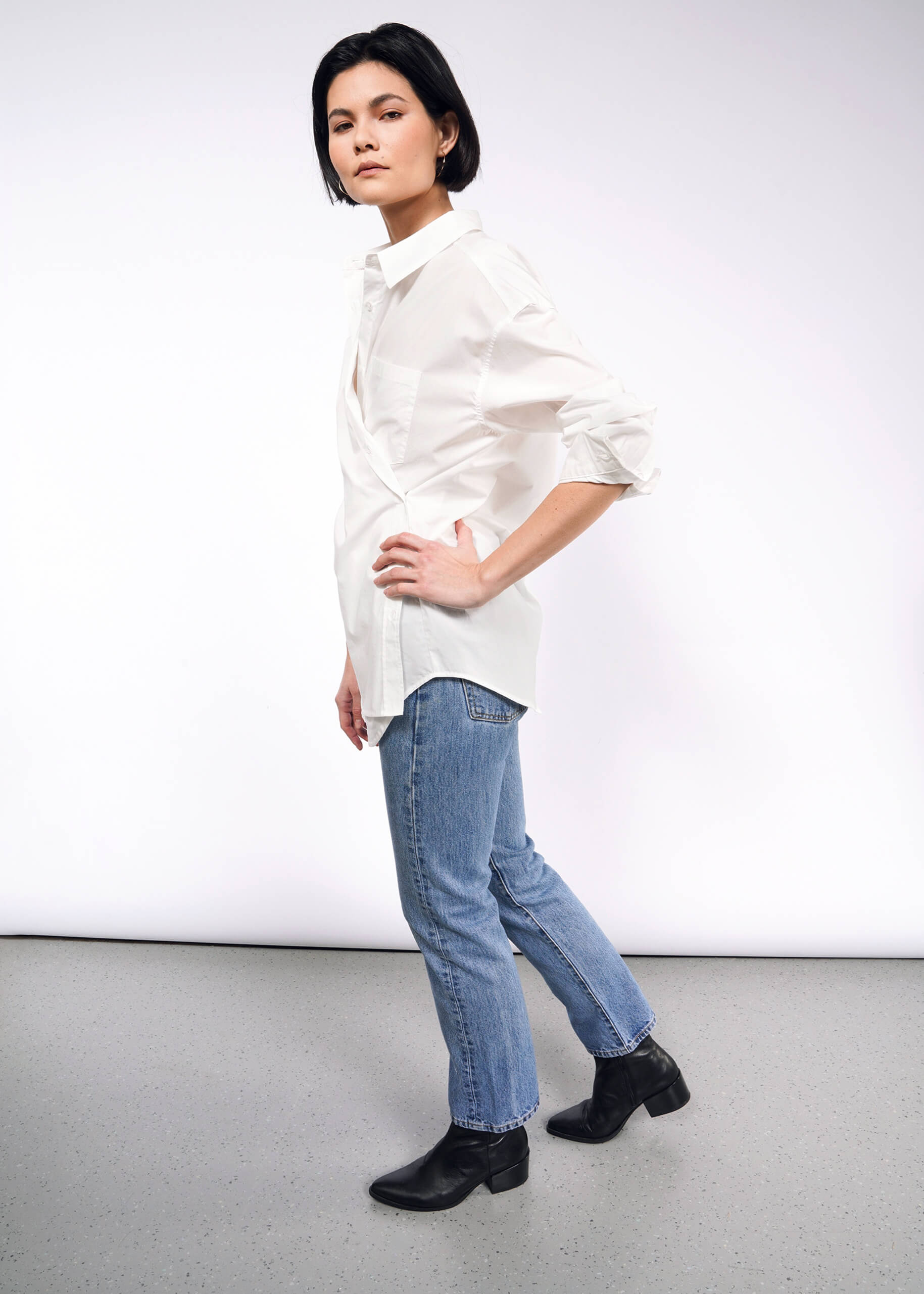 Against a plain white background, a person with short dark hair stands confidently, wearing The Essential Long Sleeve Oversized Wrap Button Up shirt with blue jeans. They pair this look with black ankle boots and have one hand on their hip, enhancing the outfits sophistication with French cuffs.