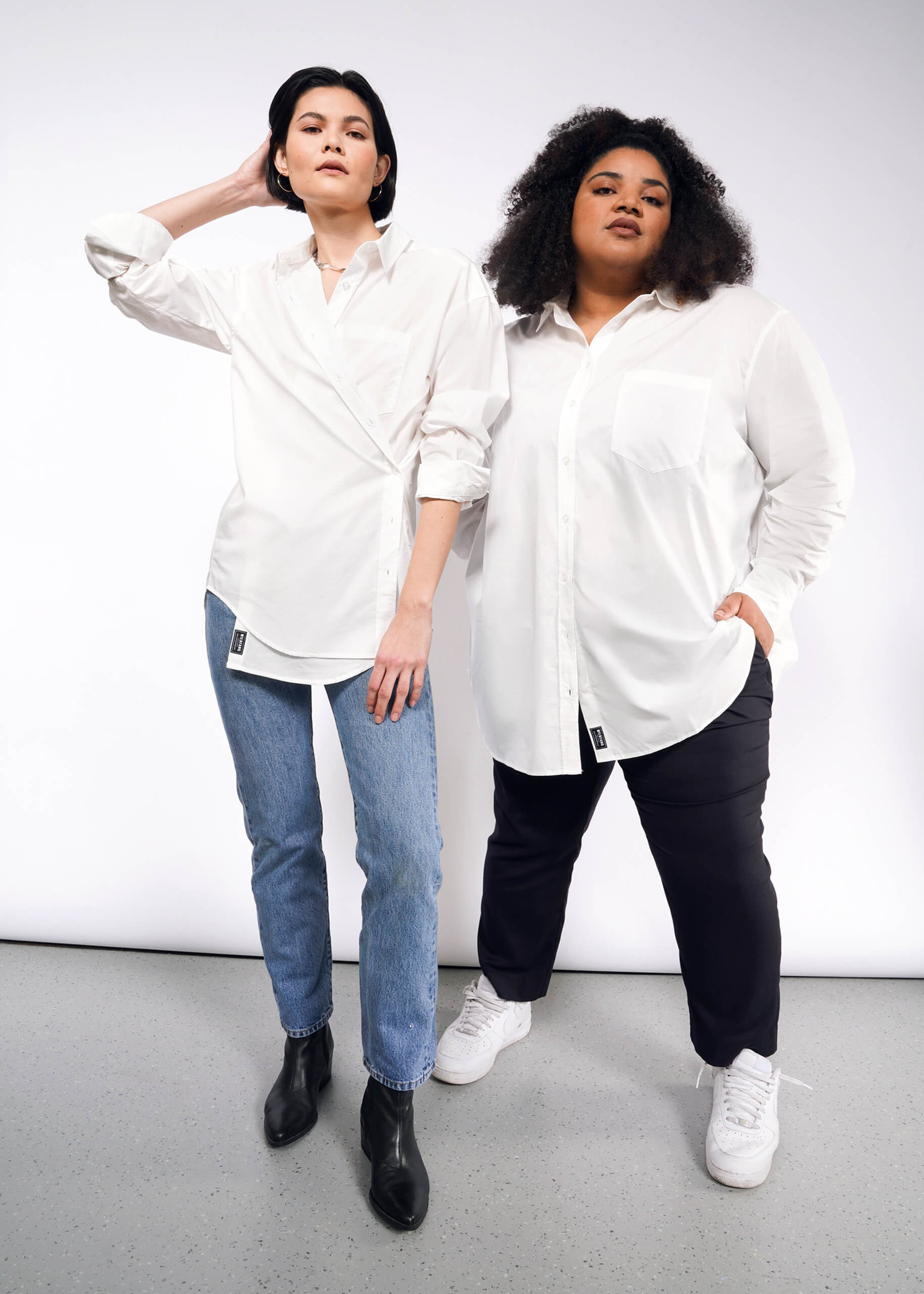 Two people stand confidently against a plain backdrop. Both wear The Essential Long Sleeve Oversized Wrap Button Up in white; one pairs it with blue jeans and black boots with French cuffs, the other with dark pants and white sneakers. The person on the left poses with a hand on their head.