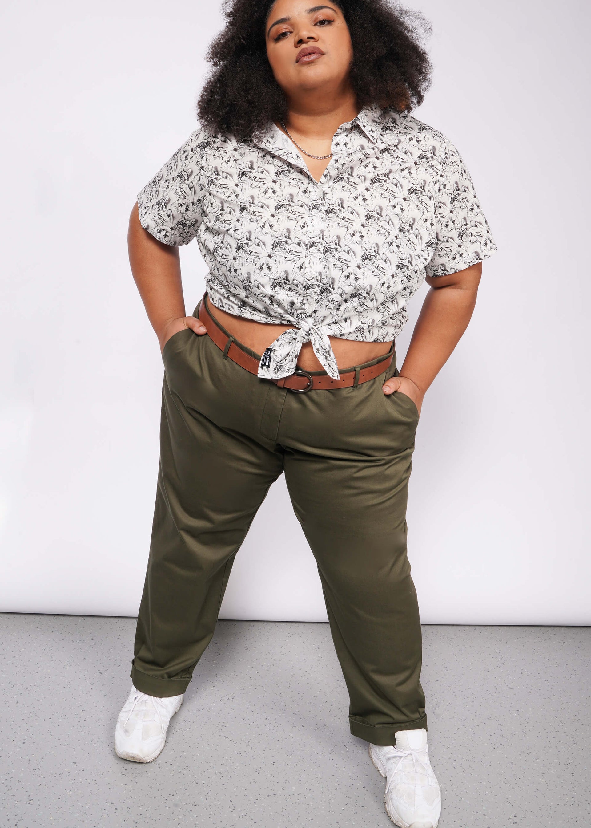 A curly-haired person confidently stands with hands in pockets, wearing a patterned short-sleeve shirt tied at the waist. They pair The Essential Trouser in olive green, a wardrobe staple, with a brown belt and white sneakers against a gray and white gradient background.