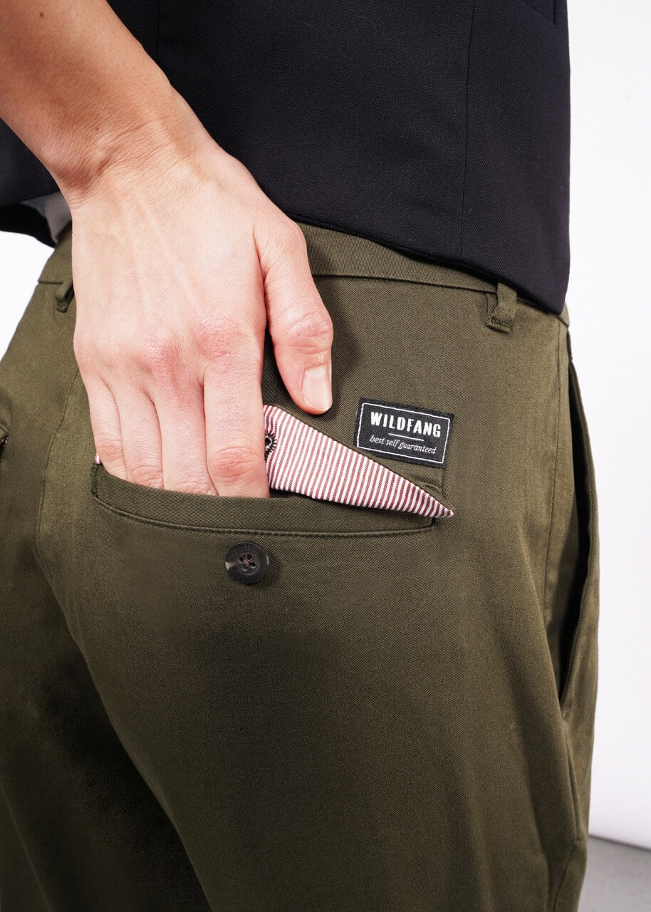 A person in olive green Essential Trousers and a dark top puts a striped cloth into the back pocket. These trousers, a wardrobe staple, showcase WILDFANG above the pocket.