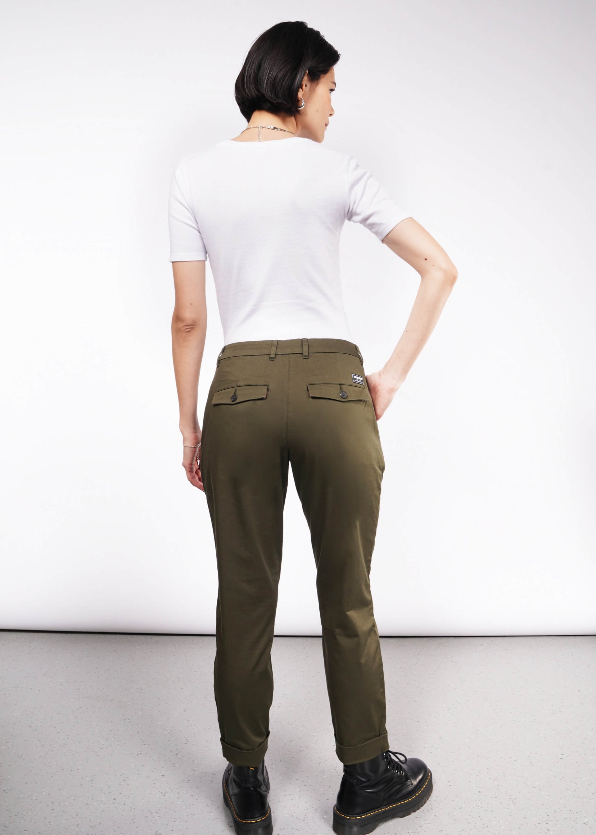 Rear view of a person with short dark hair against a white background, wearing a wardrobe staple: a white short-sleeved shirt, The Essential Trouser in classic olive green, and black shoes. They face away with one hand on their hip.