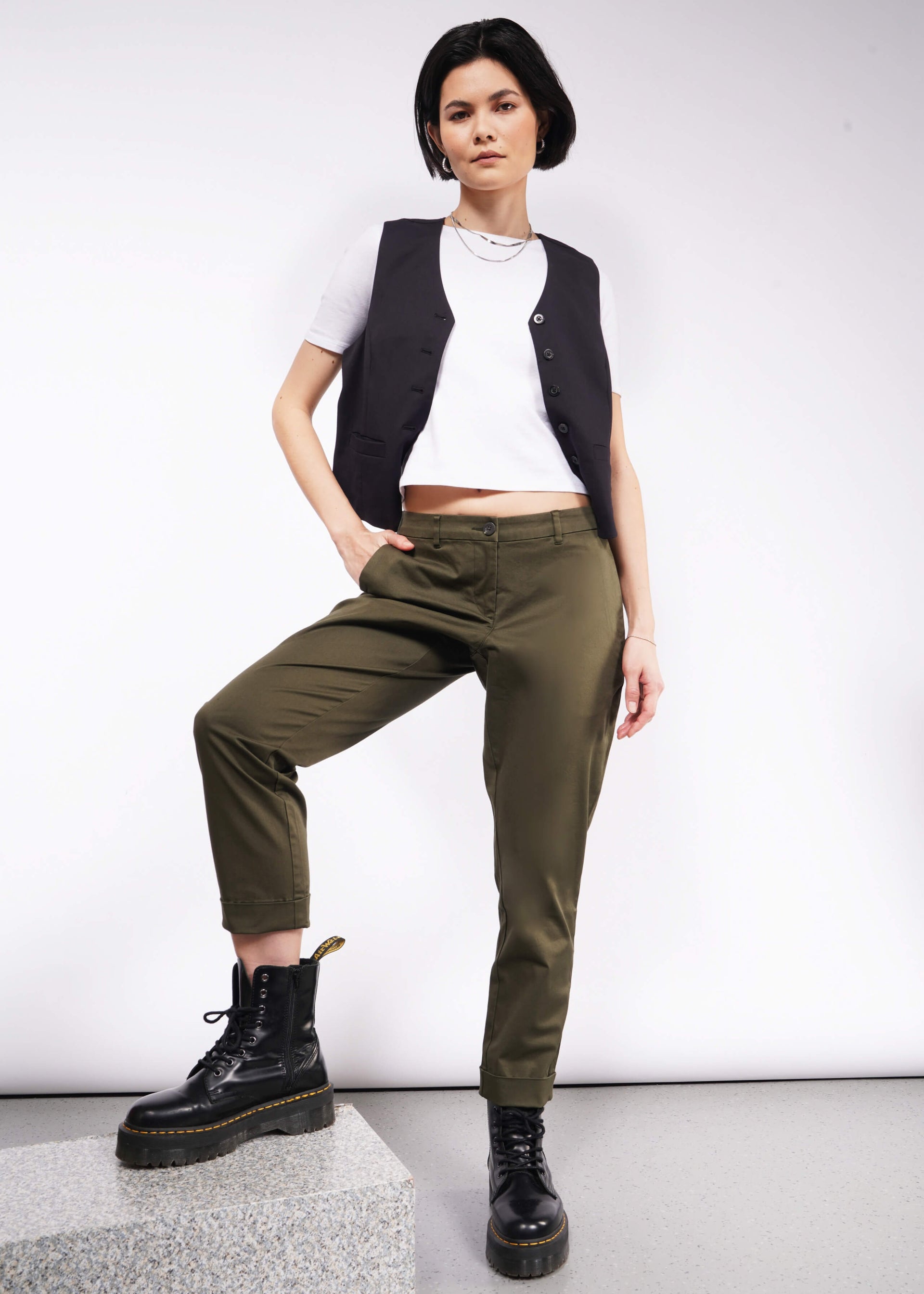 A person with short dark hair poses confidently in a white t-shirt, a black vest, and The Essential Trouser in olive green. These wardrobe staples pair seamlessly with their black boots as they stand with one hand in their pocket against a neutral background.