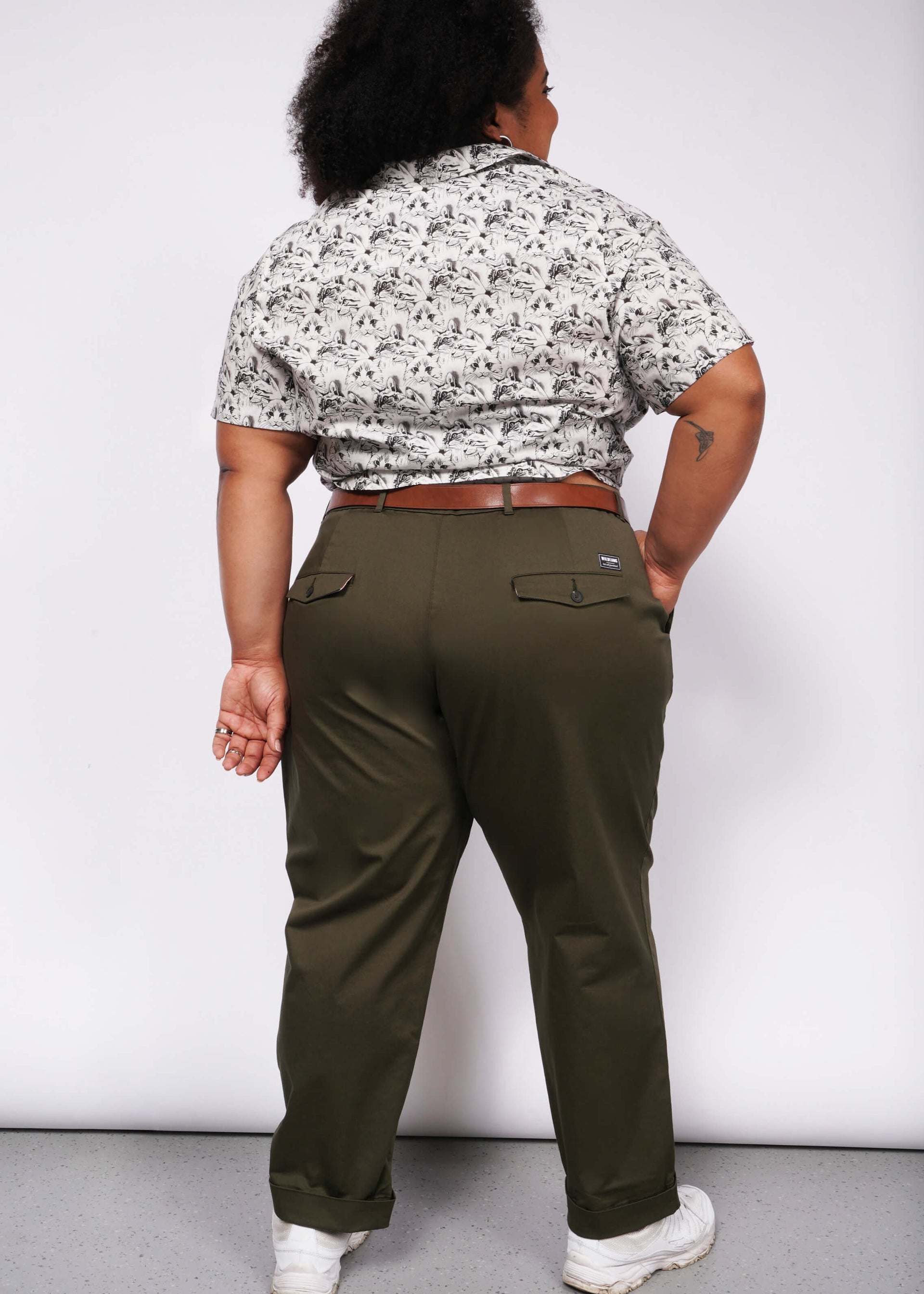 Against a white backdrop, a person faces away, wearing a patterned shirt with convertible cuffs and The Essential Trouser in olive green, belt-cinched as a wardrobe classic. Their feet are adorned with white sneakers, and their right forearm features an intriguing tattoo.