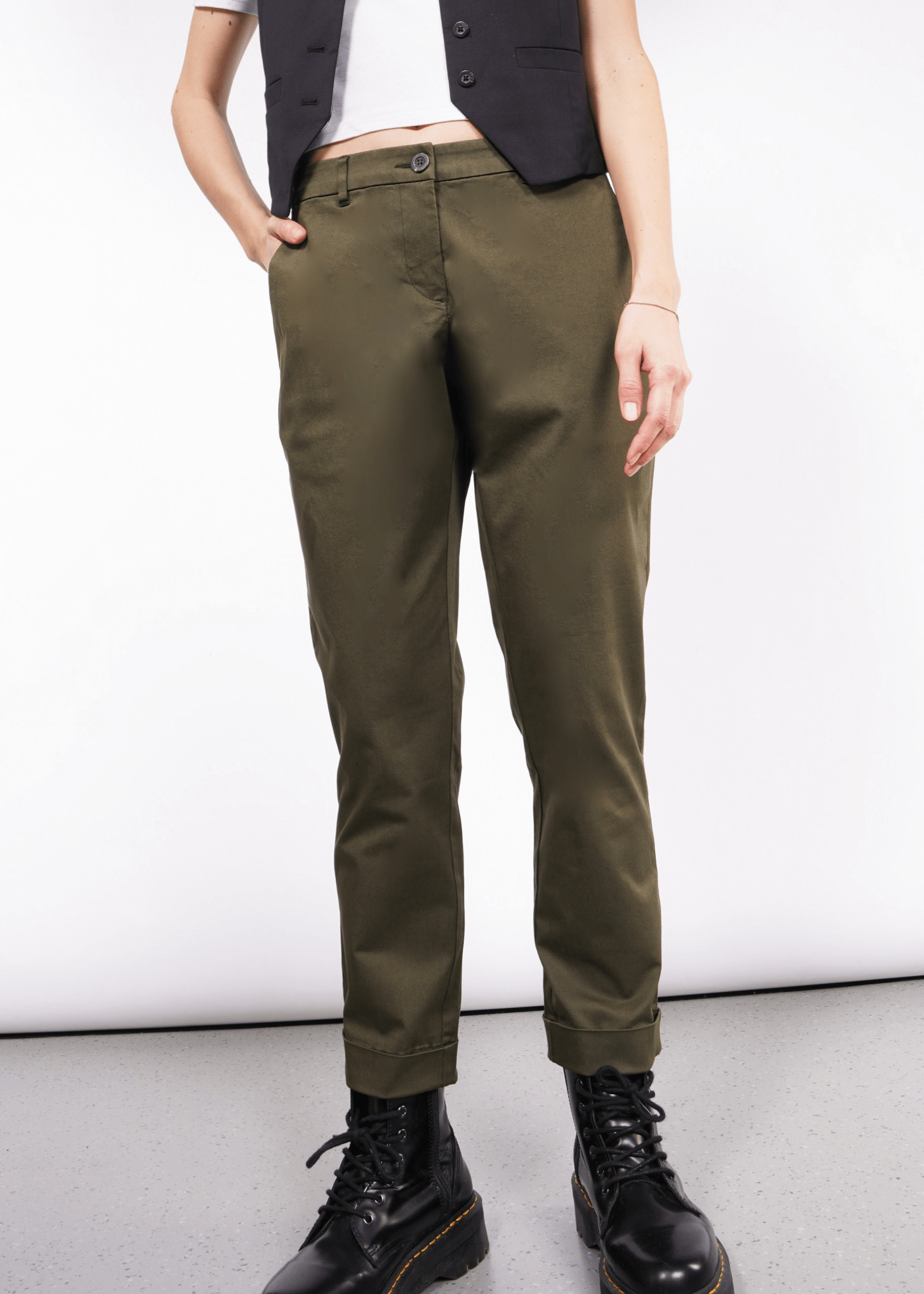 Someone wearing The Essential Trouser in olive green, perfectly paired with a black sleeveless vest and sleek black boots. Their hands rest casually in the pockets against a plain white background that enhances the minimalist aesthetic.