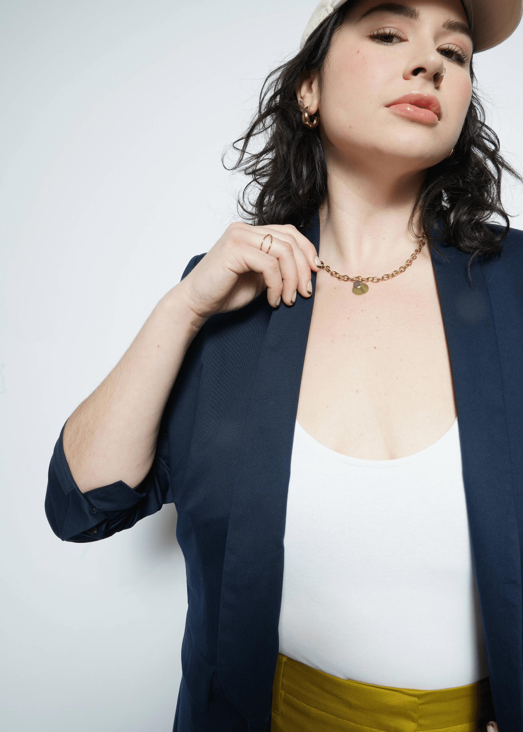 A woman in The Essential Tux Blazer with rolled-up sleeves layers it over a white top and yellow pants. Shes styled with a beige cap, gold chain necklace, and hoop earrings. Her dark curly hair and confident gaze enhance the jackets tailored fit, perfectly complementing her style.