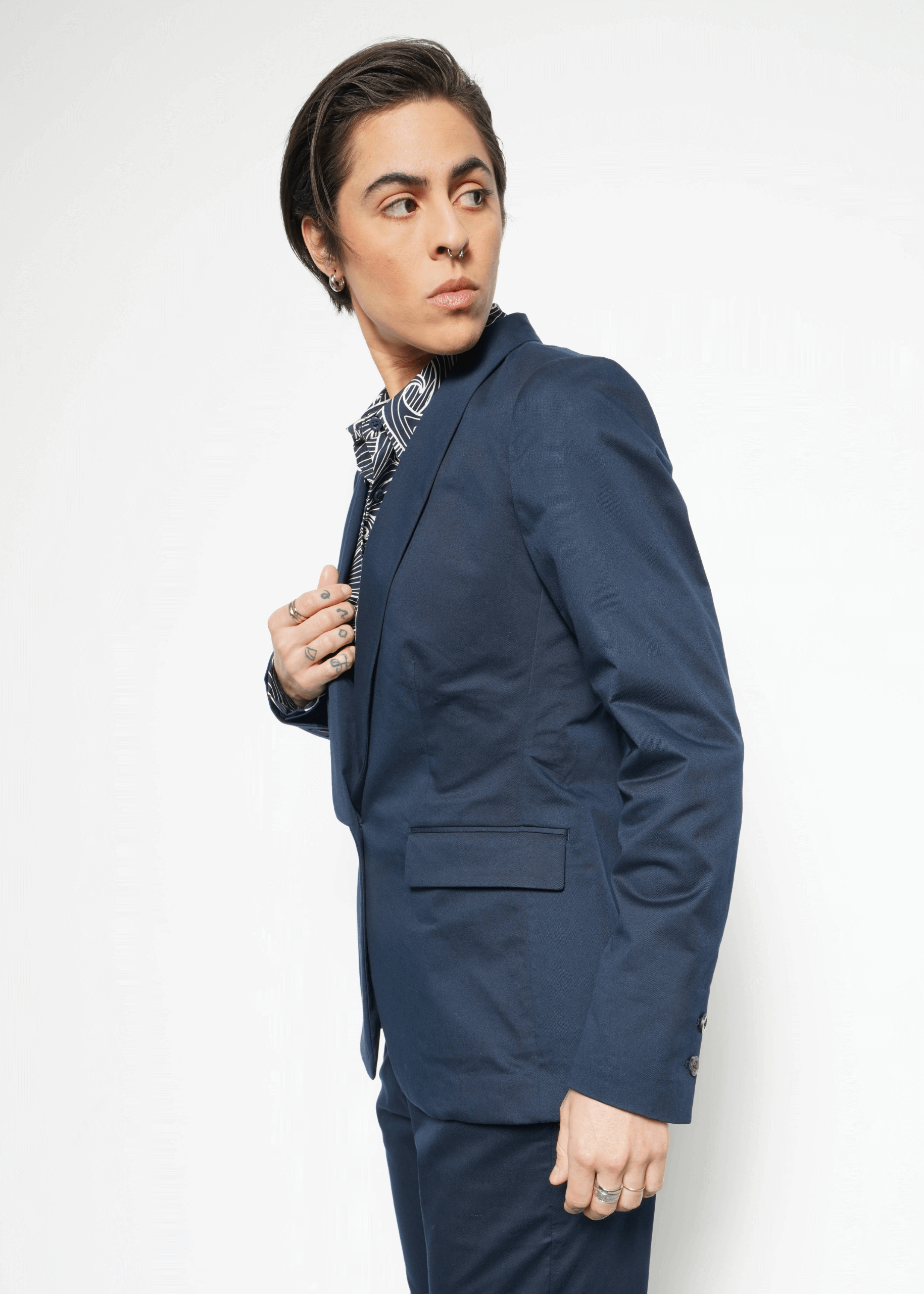 A person with short dark hair, wearing The Essential Tux Blazer and a patterned shirt, stands against a white background. The blazers tailored fit enhances their thoughtful expression as they turn slightly to the side, gazing into the distance.