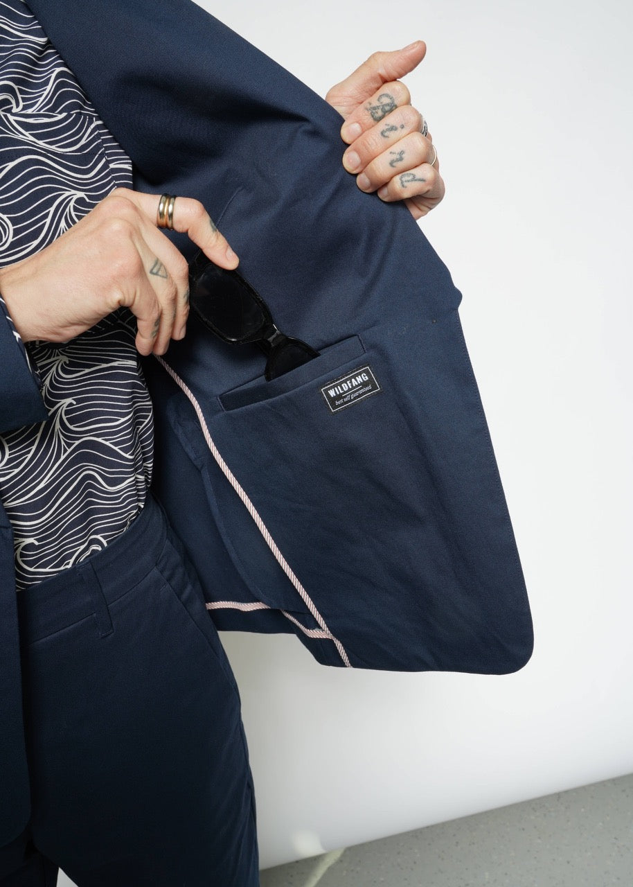 A person in a navy The Essential Tux Blazer reveals the lining and an interior pocket with a label while holding black sunglasses. Their left hand showcases tattoos and rings. The shirt beneath, made from breathable fabric, has a wavy pattern that complements the blazers tailored fit.