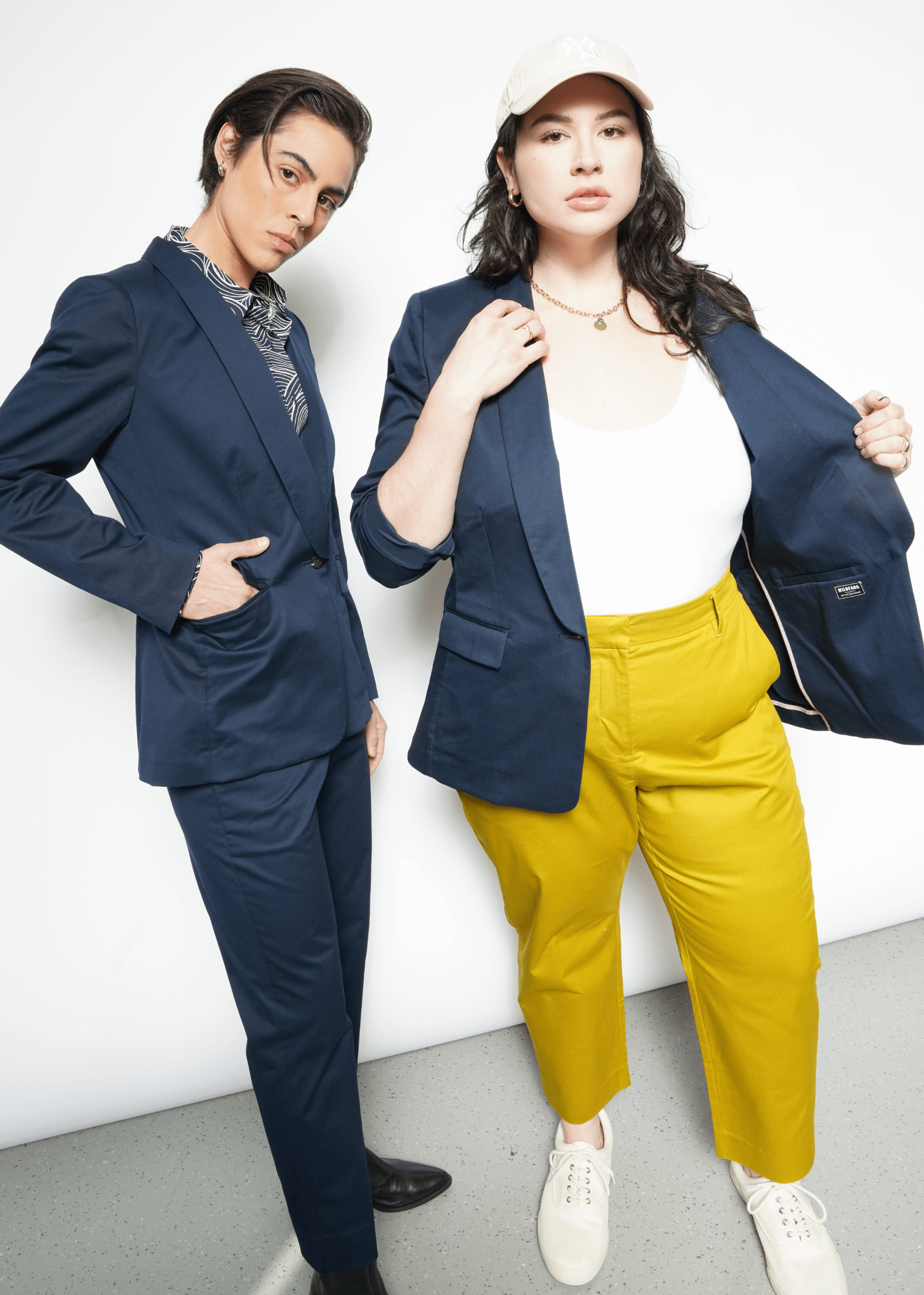 Two individuals flaunt their style in The Essential Tux Blazer. On the left, a tailored blue outfit paired with a patterned scarf complements the blazer, while on the right, its worn over a white shirt and yellow pants for contrast. Their ensembles pop against the crisp white backdrop.