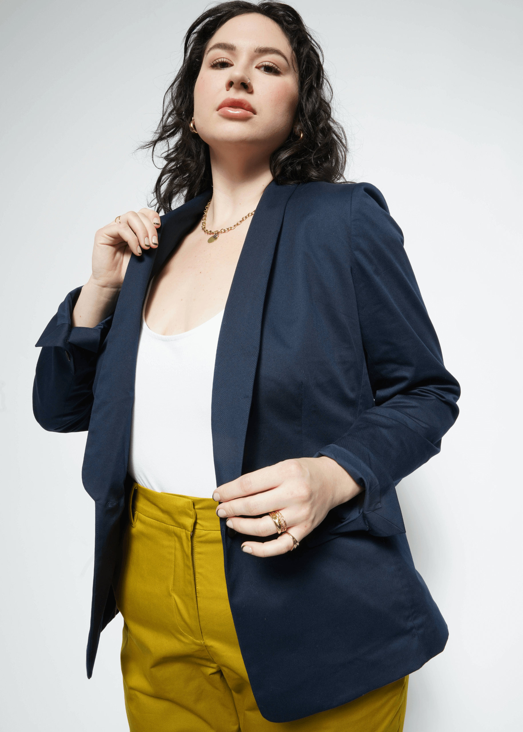 A woman exudes confidence in The Essential Tux Blazer (navy) over a white top and yellow pants. Her tailored blazer pairs with a necklace and rings, as she holds the lapel against a plain background, highlighting the sophistication of the breathable fabric.