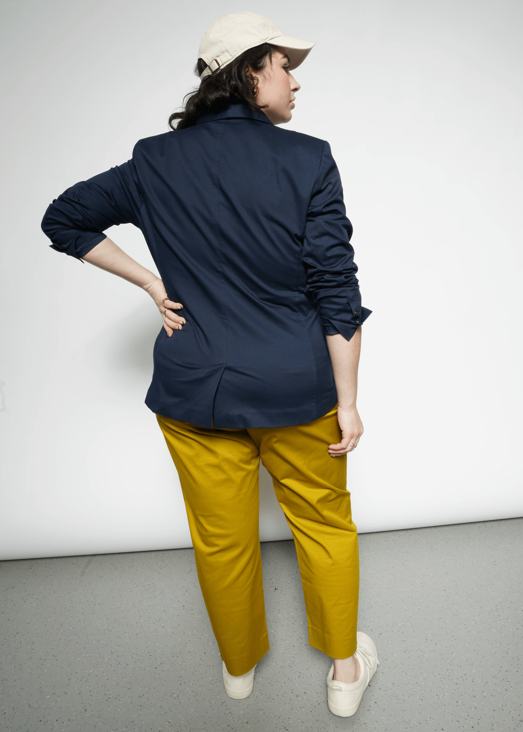 Wearing The Essential Tux Blazer and mustard pants, a person stands with their back to the camera, hand on hip. A beige cap and light shoes complete the tailored look, presenting a stylish silhouette against the plain white backdrop.